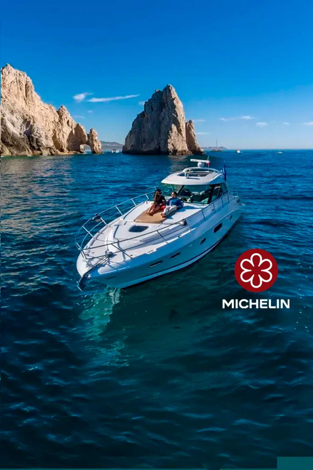  A luxury yacht cruises near the iconic rock formations of Los Cabos under a clear blue sky, with two people relaxing on deck