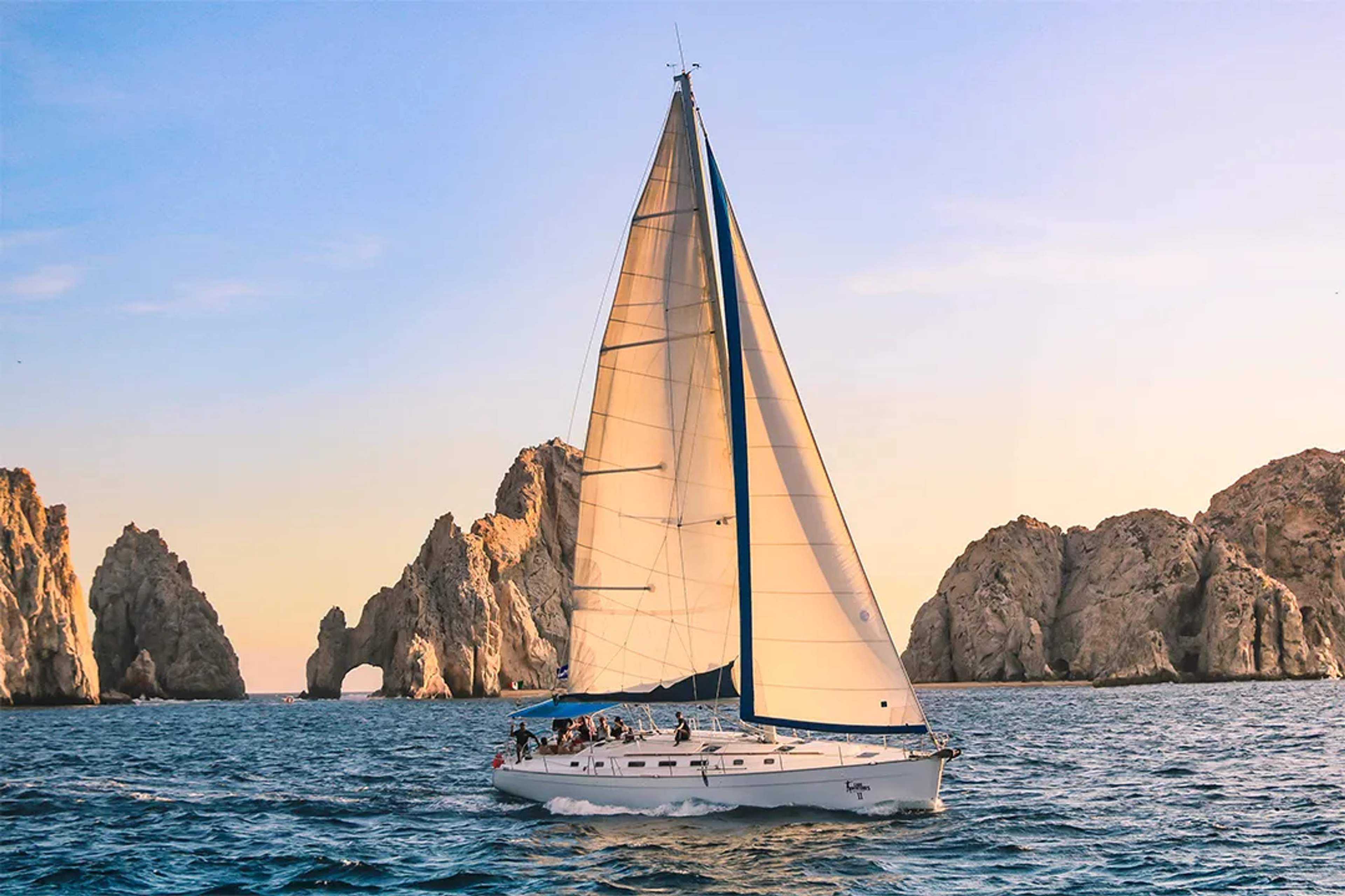 Sailboat cruising at sunset near the iconic Arch of Cabo San Lucas, with golden skies and blue sea.