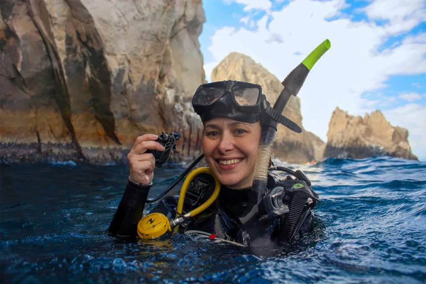 Scuba Diver In Full Gear Holding A Small Object In The Water, With Rocky Cliffs And A Partly Cloudy Sky In The Background