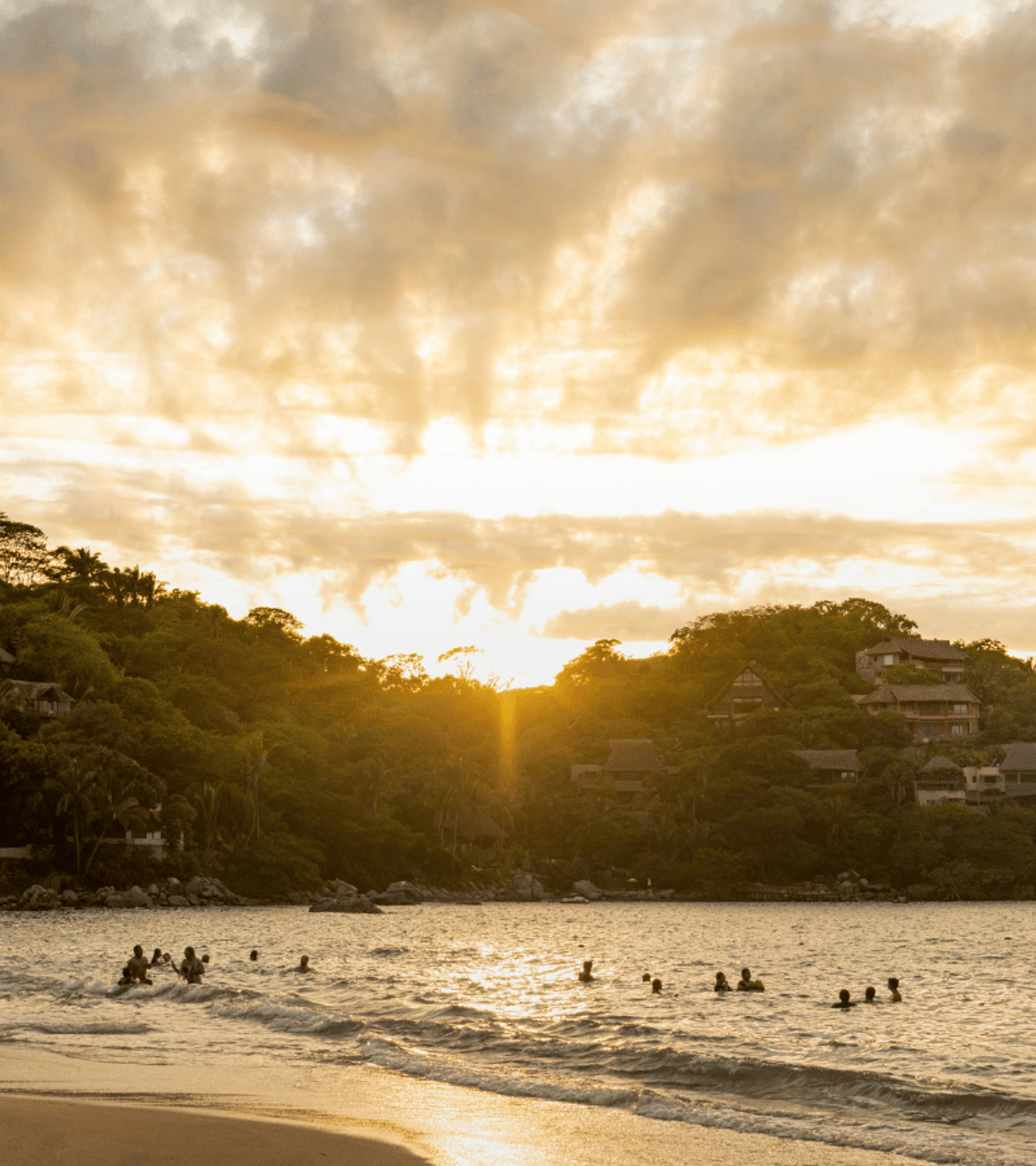 Personas nadando en el mar cerca de una playa al atardecer, con casas en colinas arboladas y un cielo parcialmente nublado.