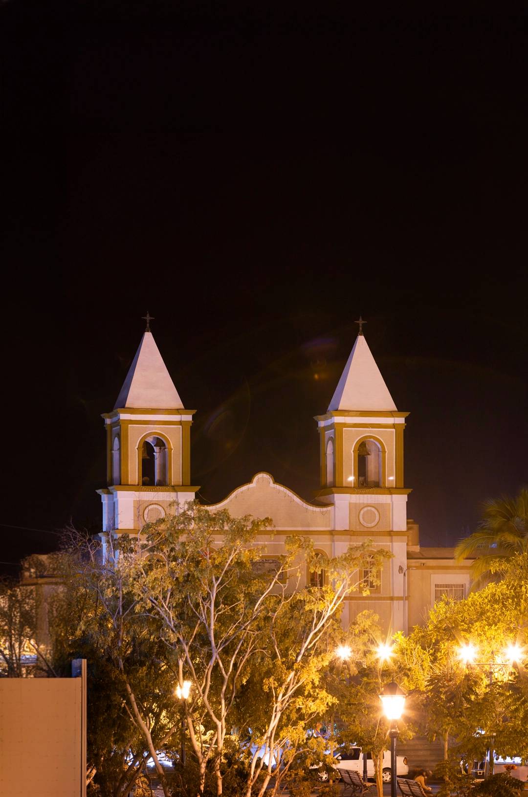 A historic church with twin bell towers illuminated at night, surrounded by trees and streetlights in San José del Cabo.