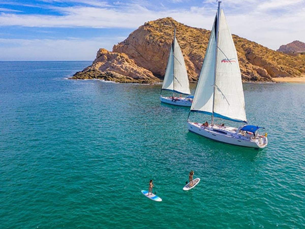 Vista aérea de dos veleros deslizándose sobre aguas tranquilas y claras de color azul cerca de una costa rocosa. Dos personas están practicando paddleboard cerca, disfrutando del día soleado y del paisaje escénico. El fondo presenta colinas escarpadas bajo un cielo brillante y parcialmente nublado.