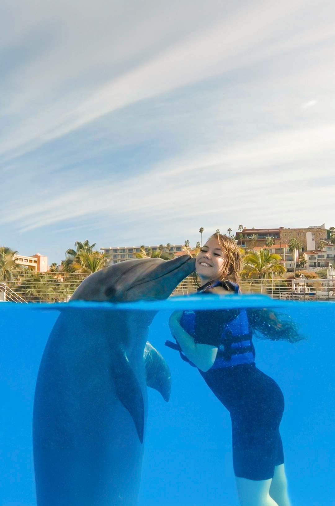 Mujer sonríe mientras recibe un tierno beso de un delfín dentro del agua cristalina.