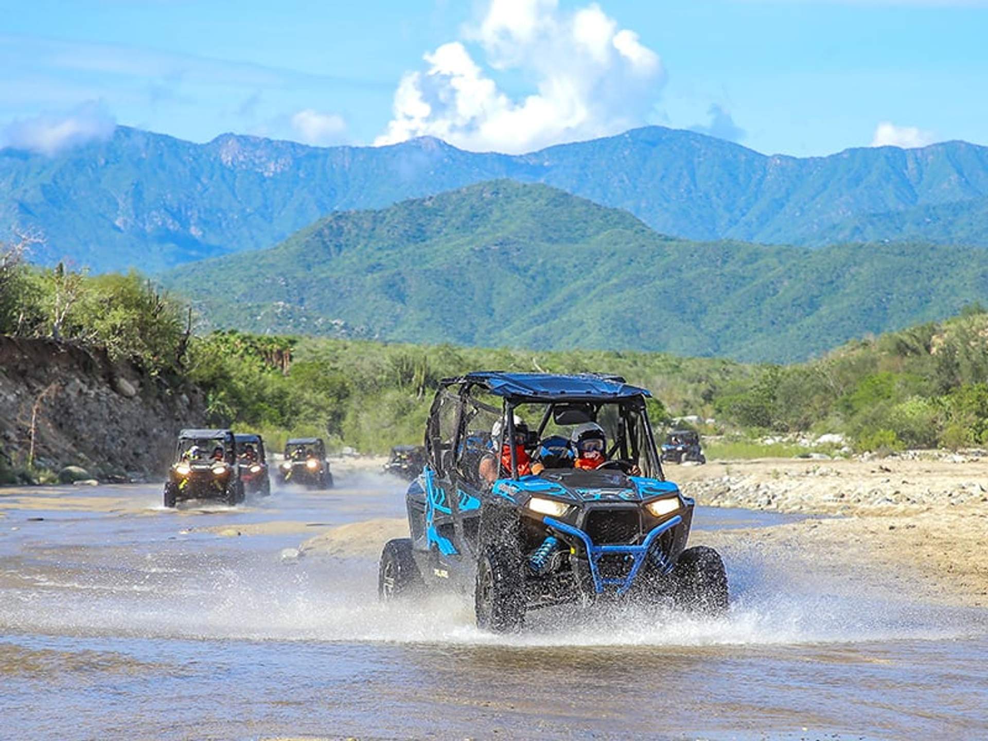 Un grupo de vehículos todoterreno cruza un río poco profundo con un paisaje montañoso de fondo.