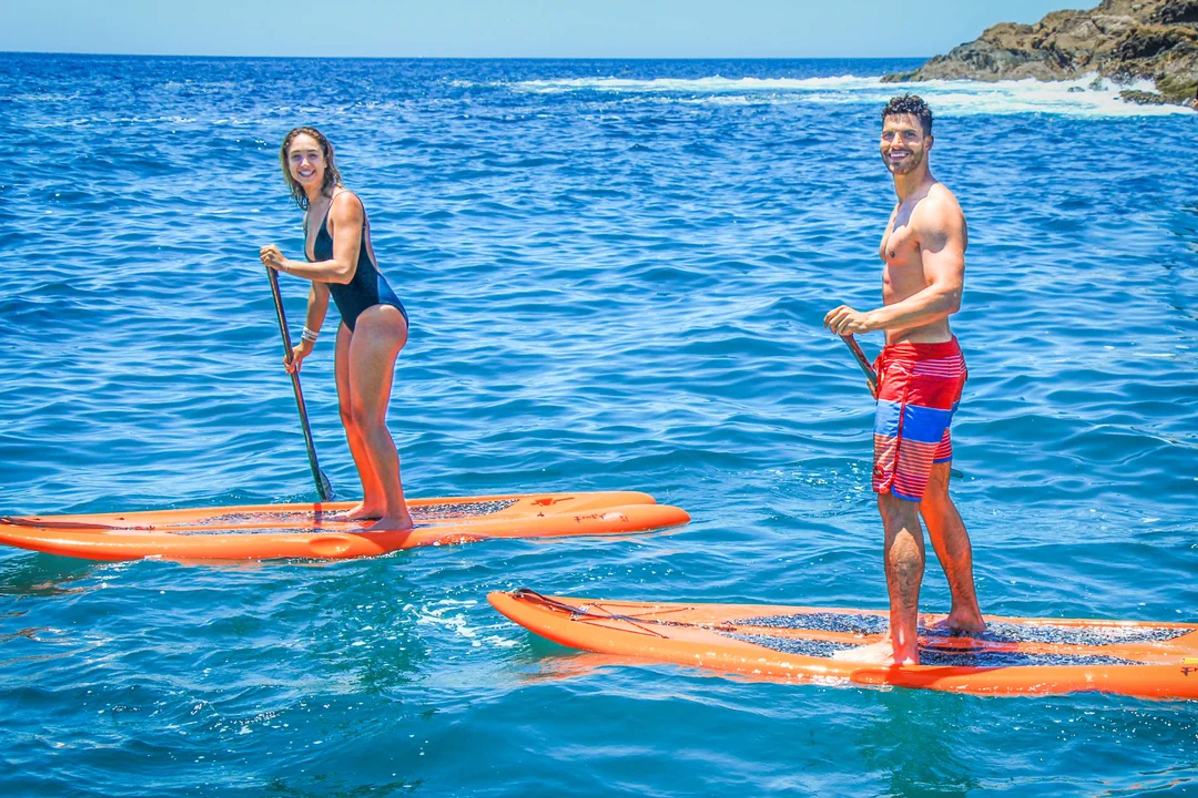 Couple paddleboarding on clear blue ocean water, smiling under the sun near rocky shores.