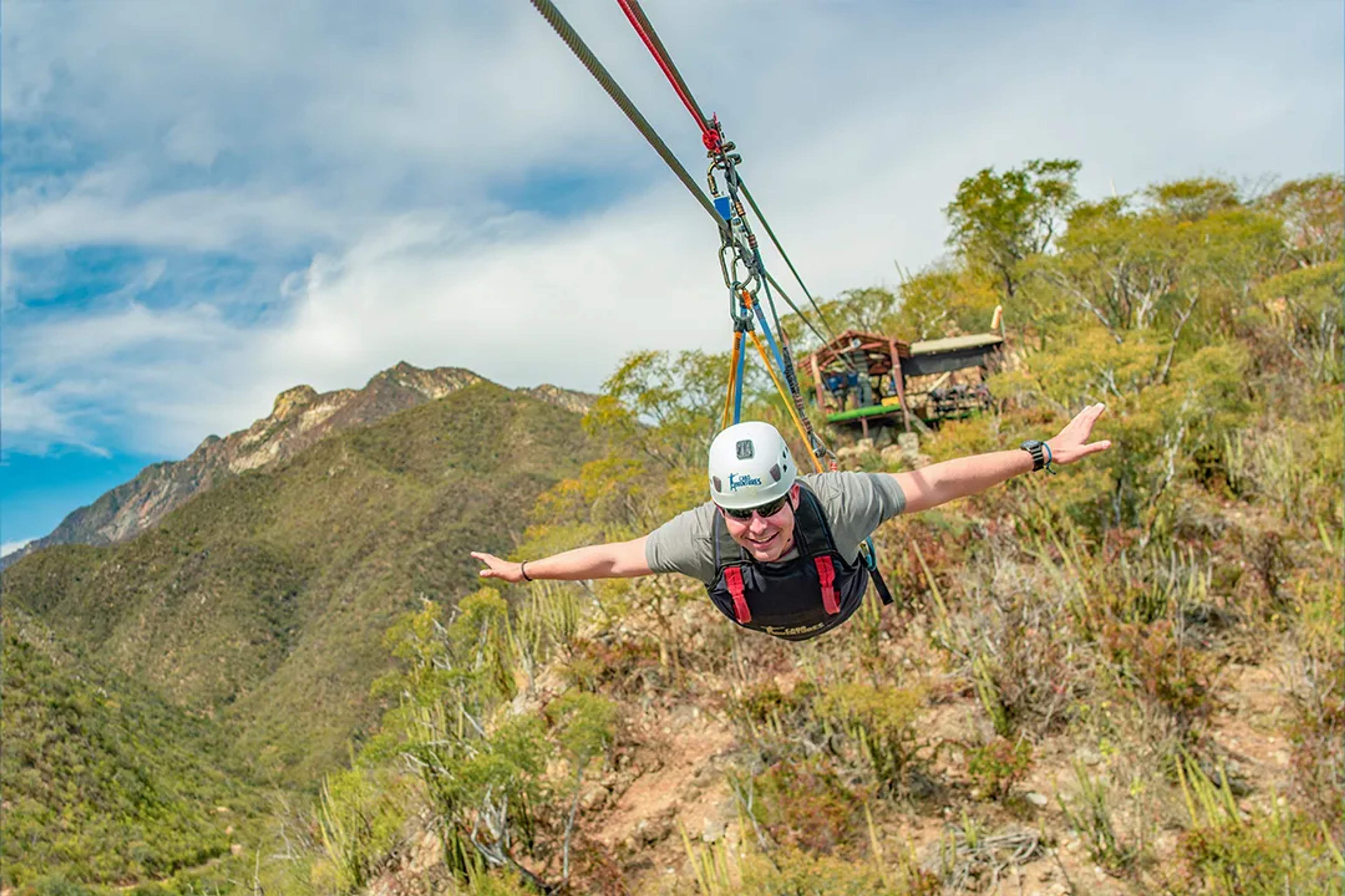 Man flying Superman-style zipline over desert canyon, arms extended, surrounded by mountains and nature