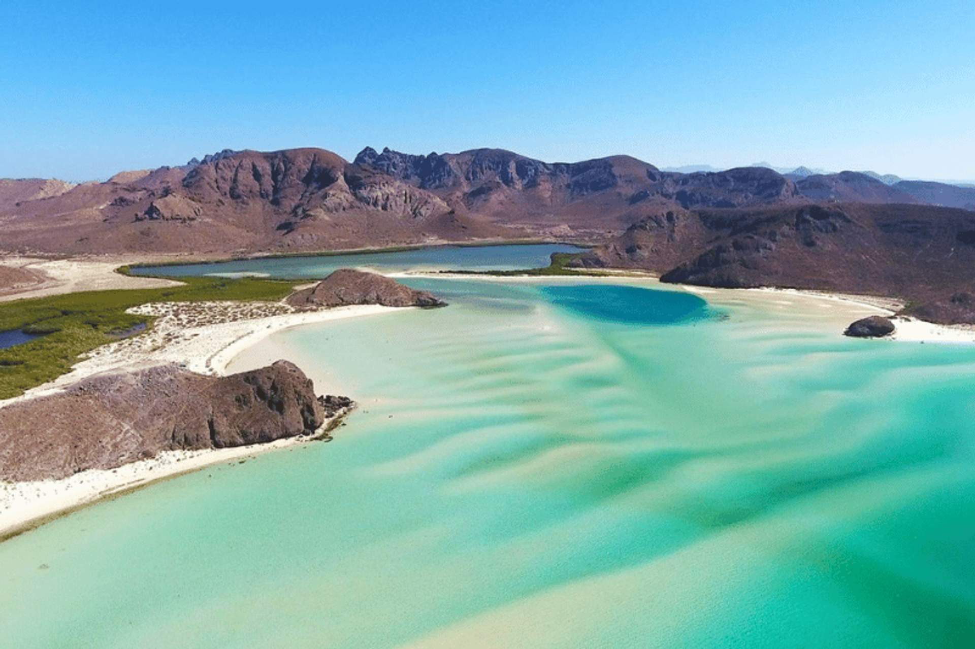 Vista aérea de una playa con aguas turquesas y arena blanca, rodeada de montañas áridas bajo un cielo despejado.