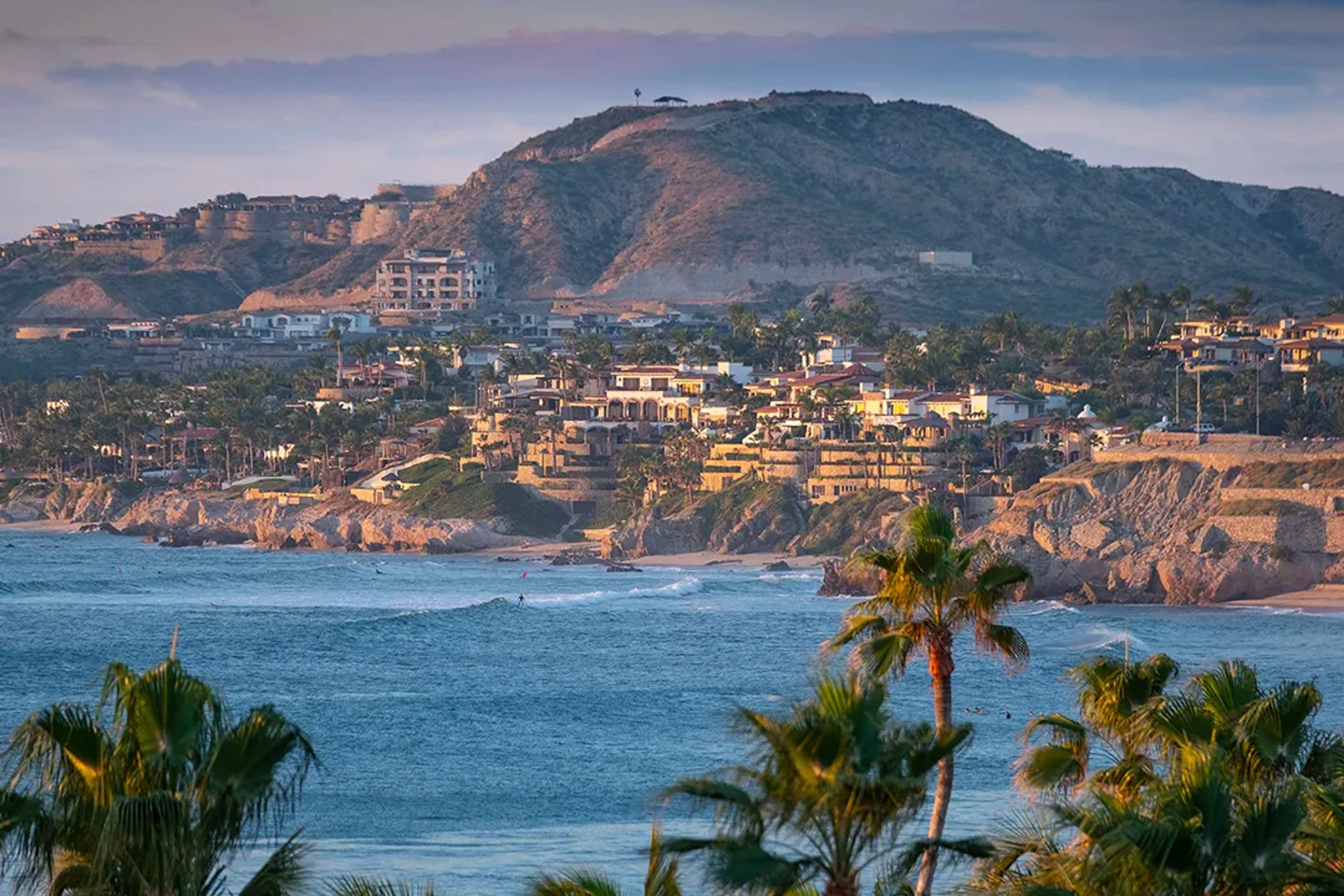 Vista panorámica de la costa de San José del Cabo con villas de lujo, acantilados y olas al atardecer.