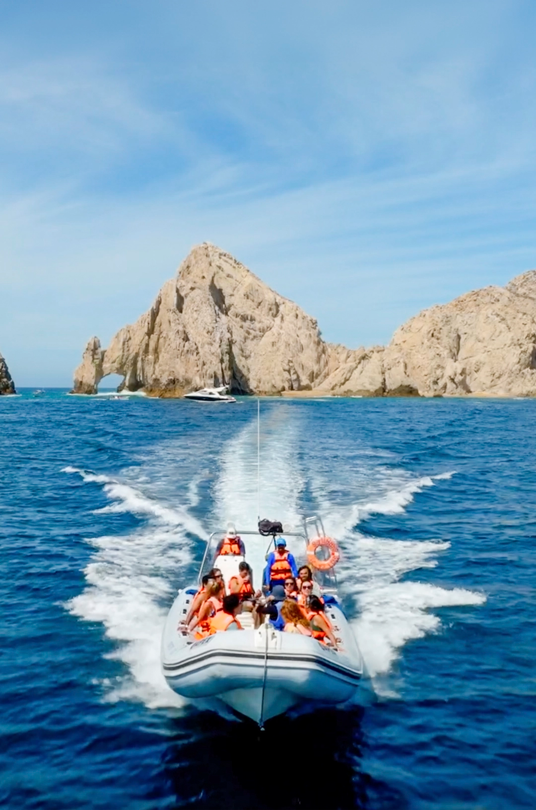 A group of people on a speedboat in the ocean with large rock formations in the background, all wearing orange life jackets.