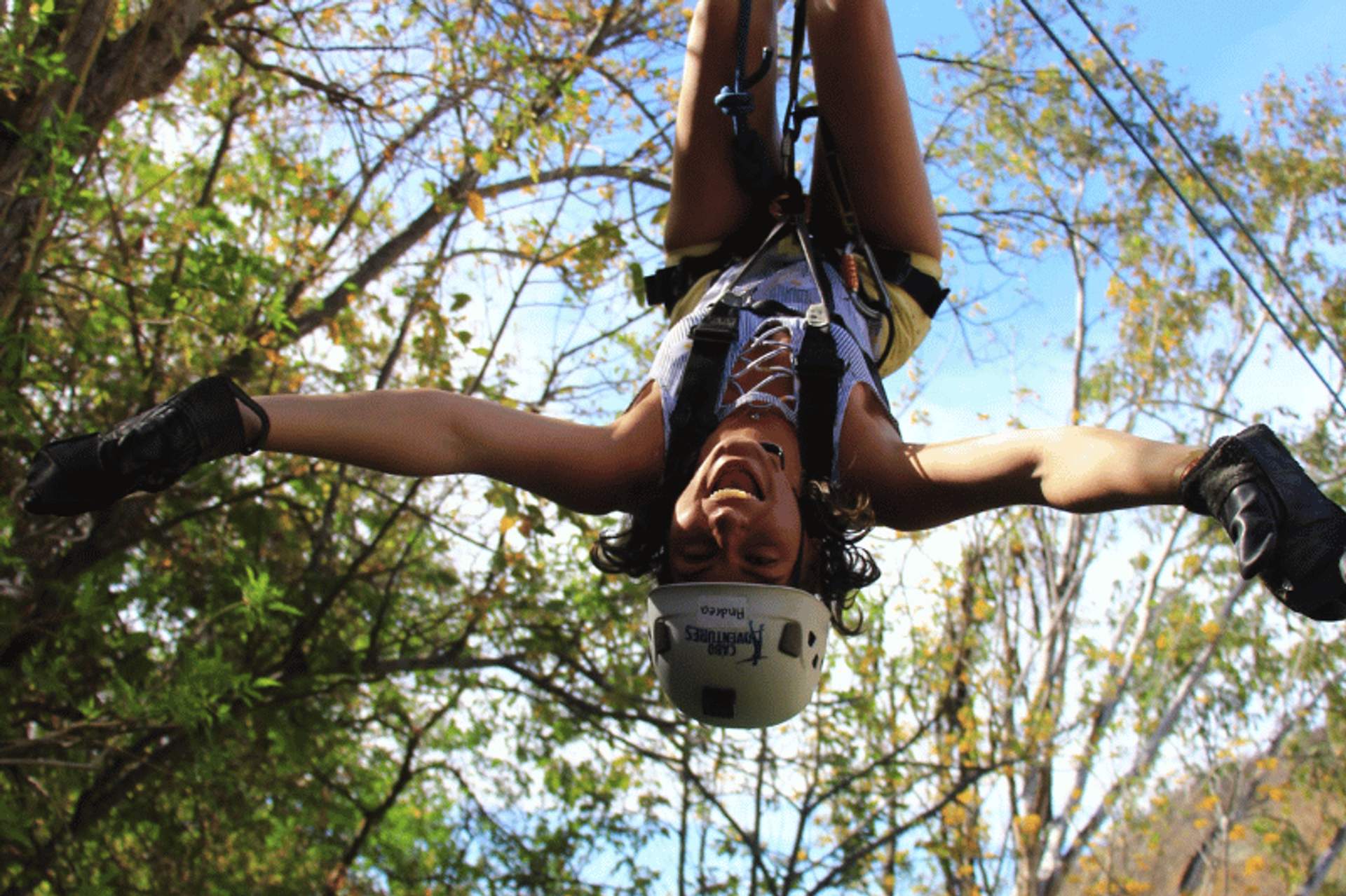 Una persona con casco y guantes cuelga boca abajo mientras se desliza por una tirolesa en un bosque, sonriendo y extendiendo los brazos.
