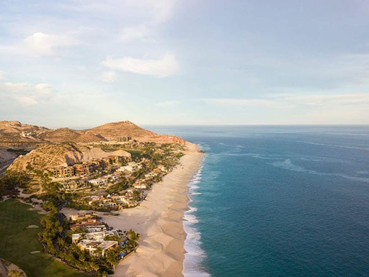 Vista aérea de un paisaje costero con una playa de arena que se extiende a lo largo del océano turquesa. La costa está salpicada de casas lujosas y exuberante vegetación, con un telón de fondo de colinas áridas bajo un cielo parcialmente nublado.
