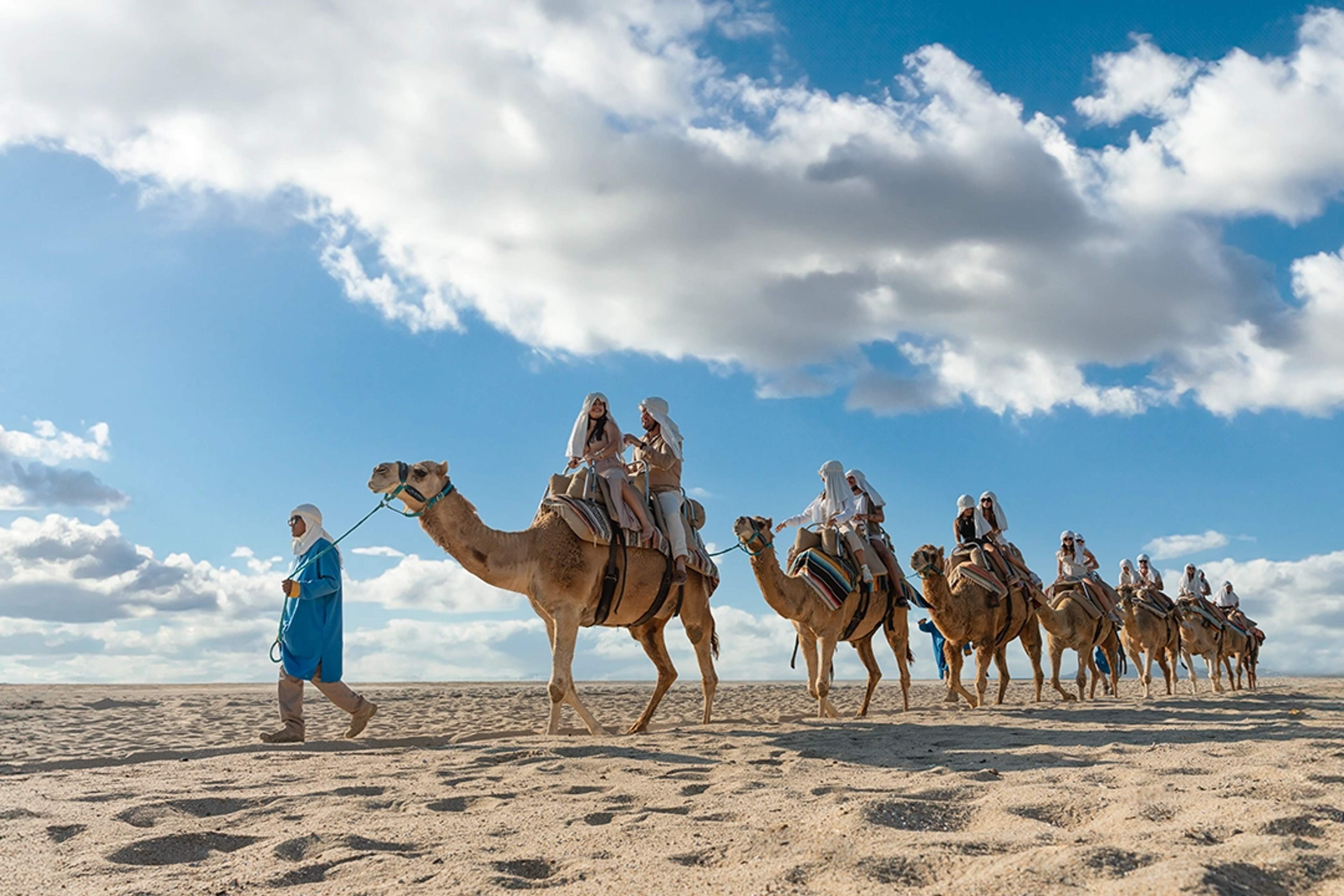 Una caravana de camellos guiada por un guía con turistas montando bajo un cielo soleado en una caminata a camello en Los Cabos.