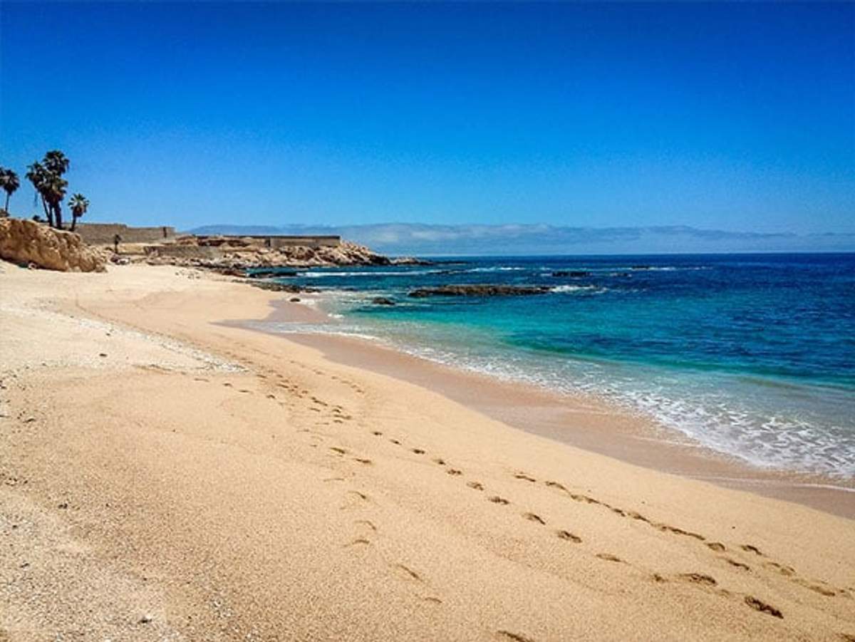 Una tranquila playa de arena con huellas claras que llevan hacia el océano turquesa. La costa está enmarcada por afloramientos rocosos y unas pocas palmeras bajo un cielo azul brillante.