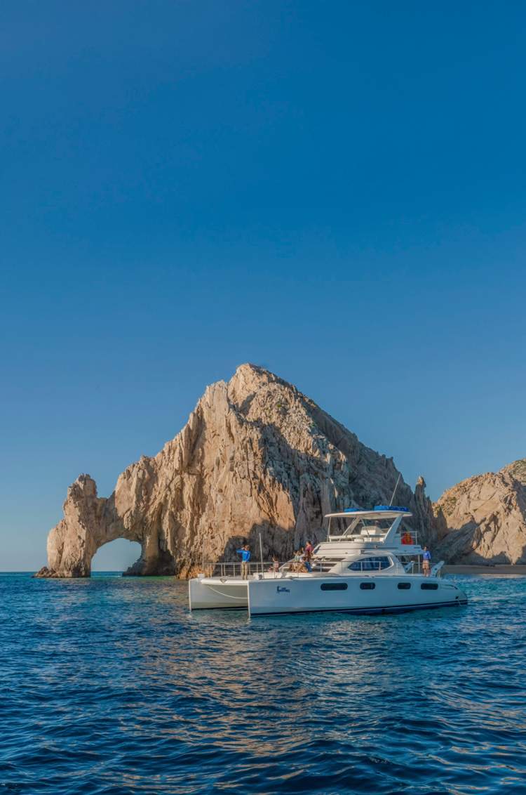 Catamarán de lujo anclado cerca del Arco de Cabo, pasajeros disfrutan de la vista icónica bajo un cielo azul claro.