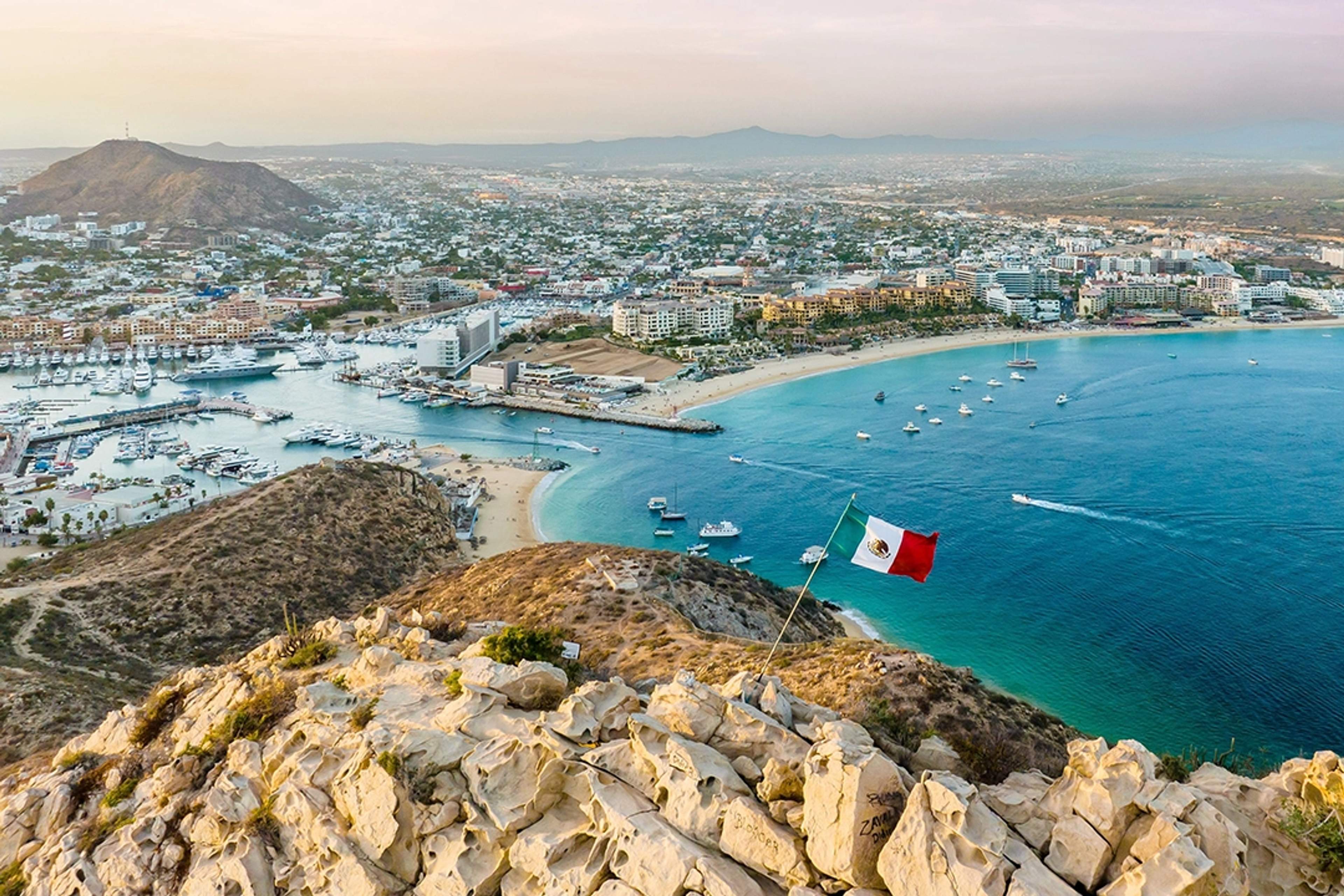 Vista panorámica de Cabo San Lucas, México, con una bandera mexicana, marina y aguas turquesas en primer plano.