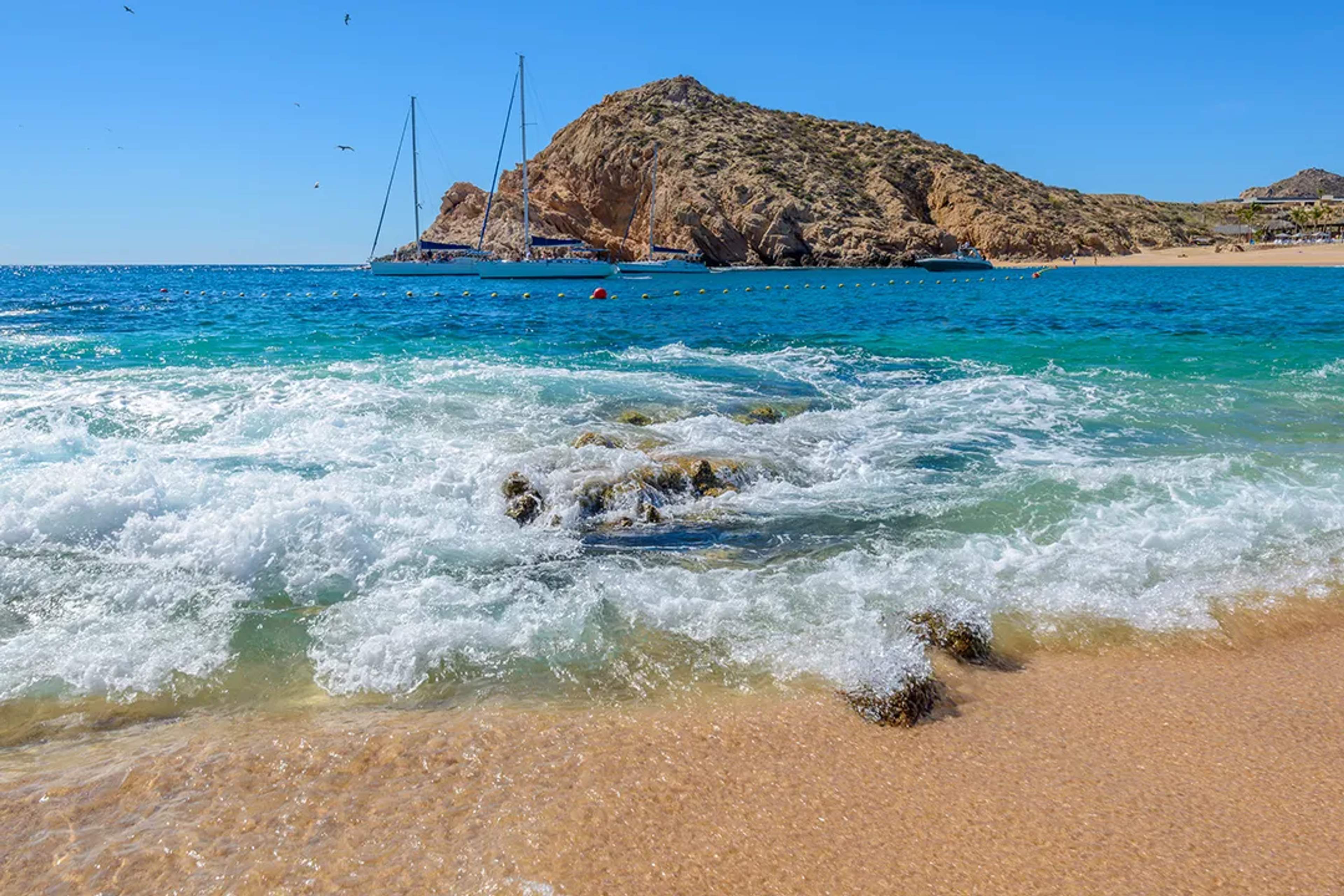 Waves crashing on a sandy beach with turquoise water and sailboats in Los Cabos.