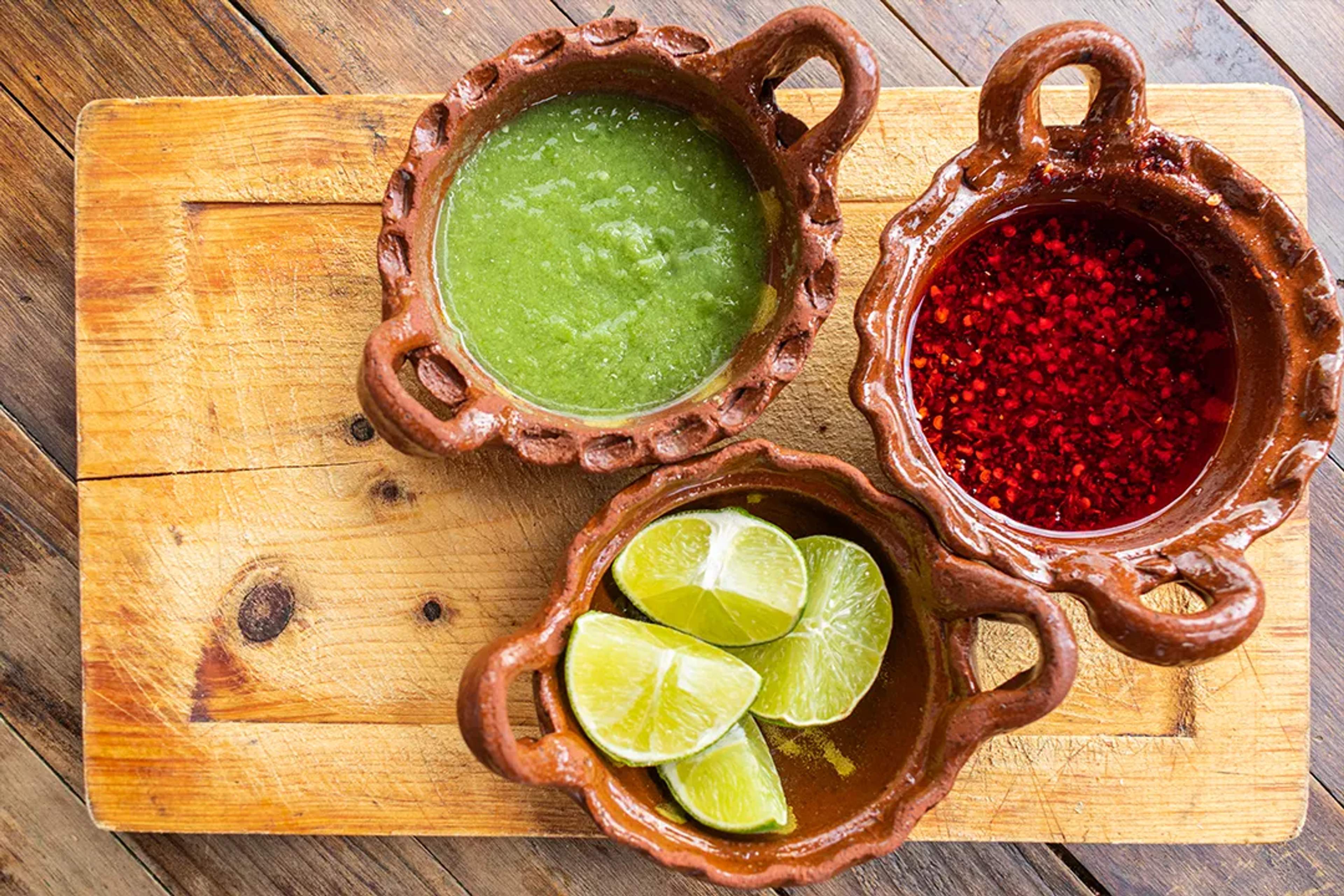 Green salsa, red chili oil, and lime wedges in clay bowls on a rustic wooden board.