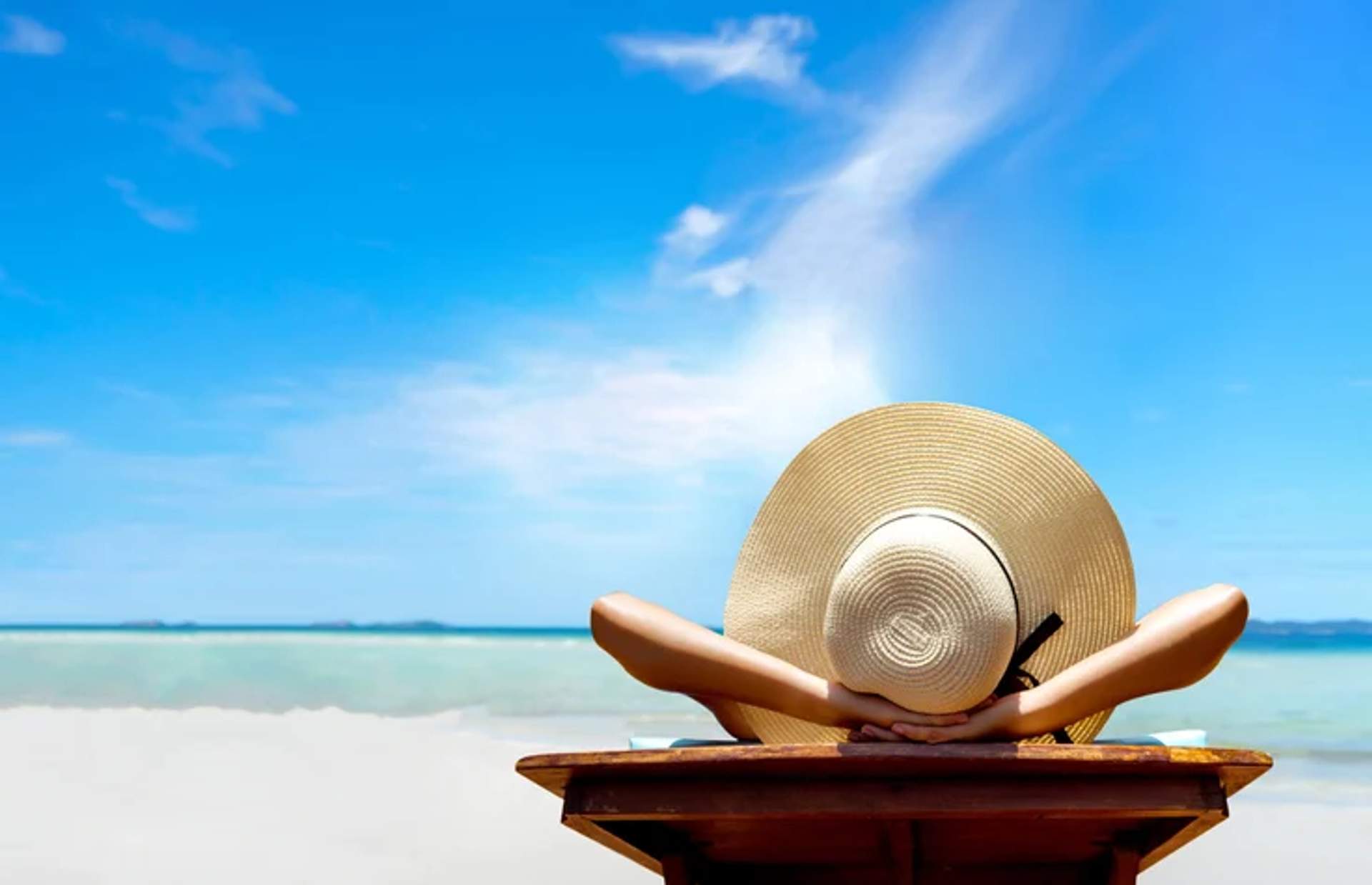Una persona con un sombrero de ala ancha se relaja en una silla de madera, frente a una playa serena con cielos azules claros y aguas turquesas.