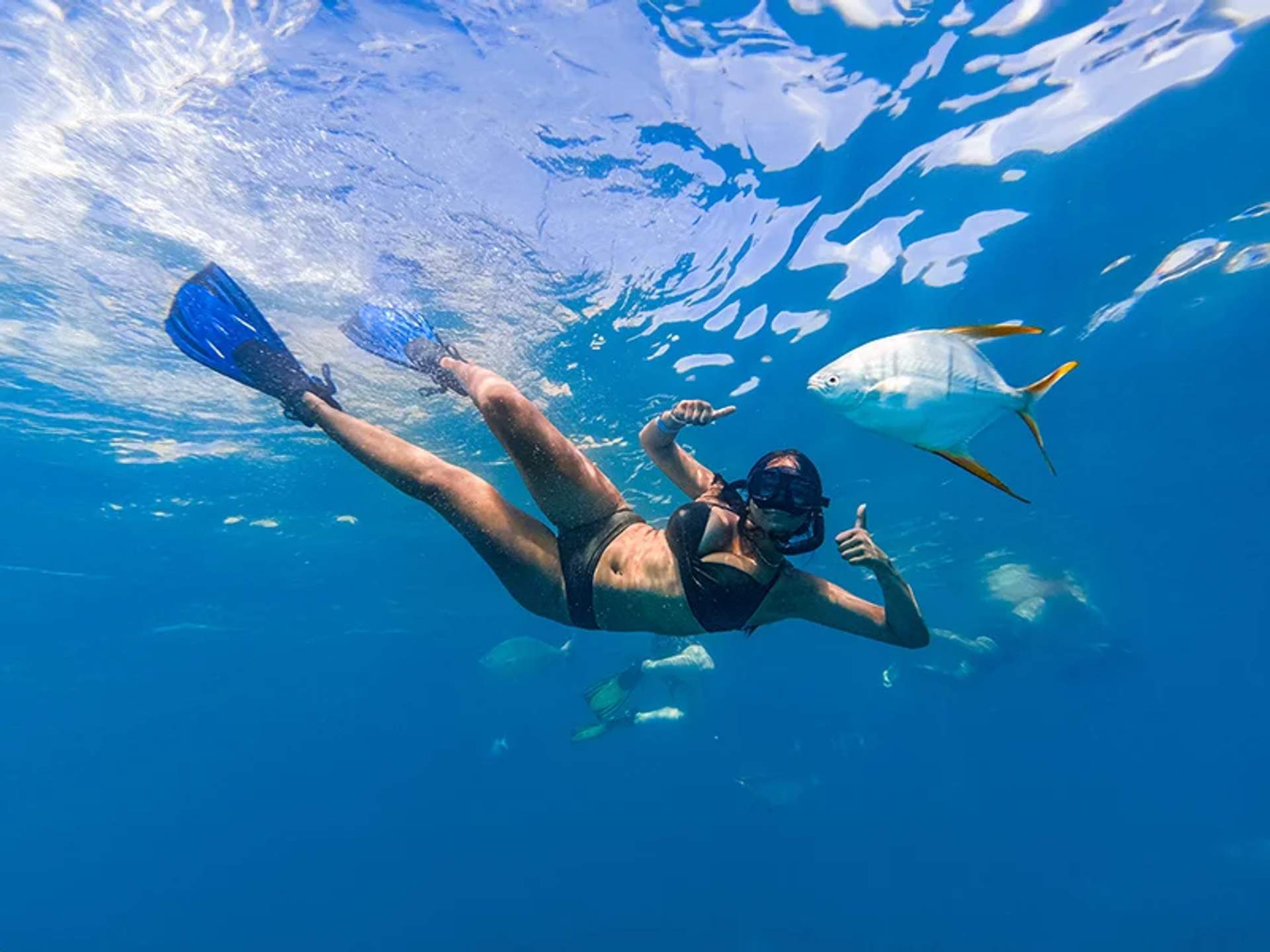 Una mujer buceando con snorkel bajo el agua hace un gesto de aprobación mientras nada cerca de un pez en aguas claras y azules.