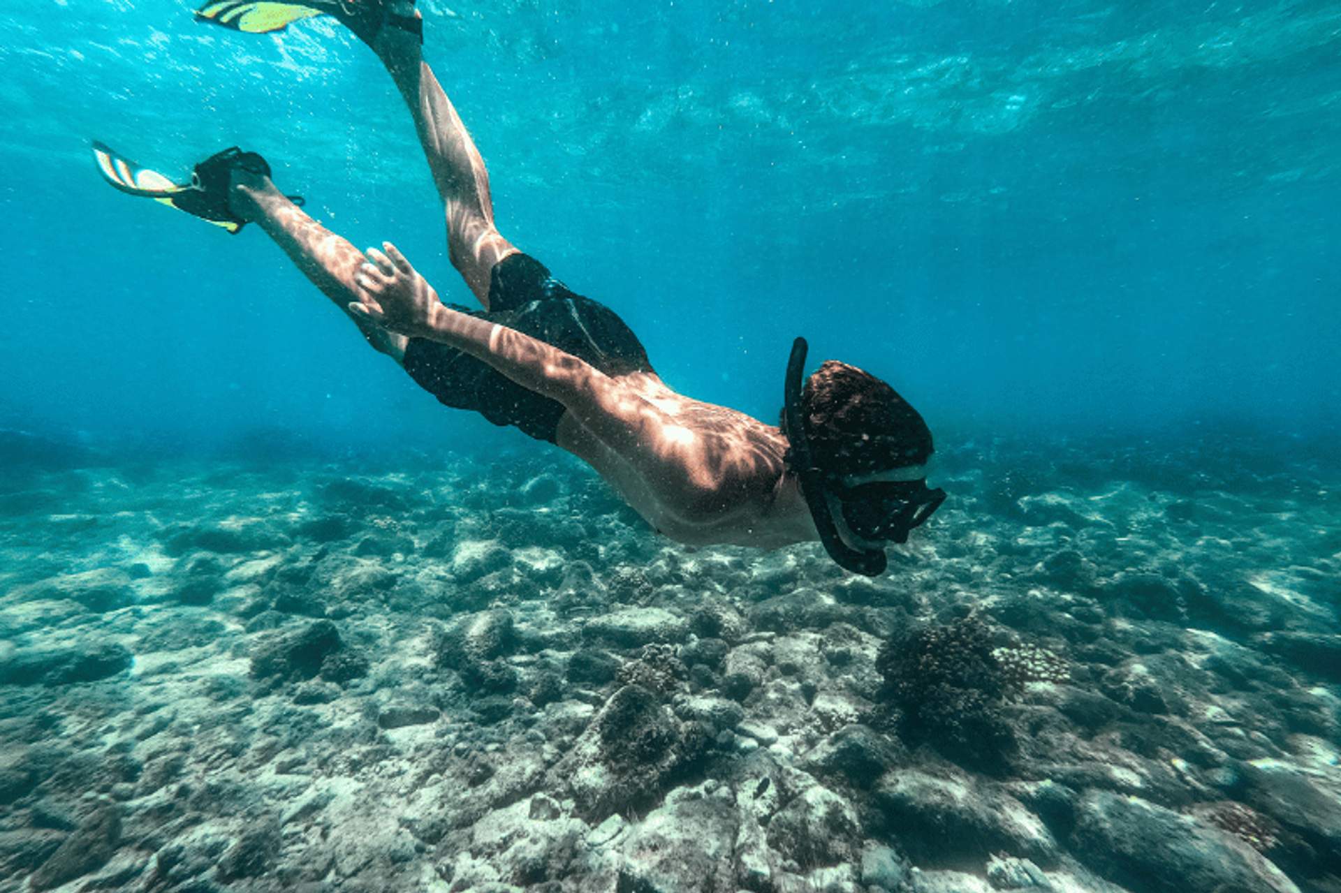 Person snorkeling underwater, exploring a rocky seabed with clear blue water.