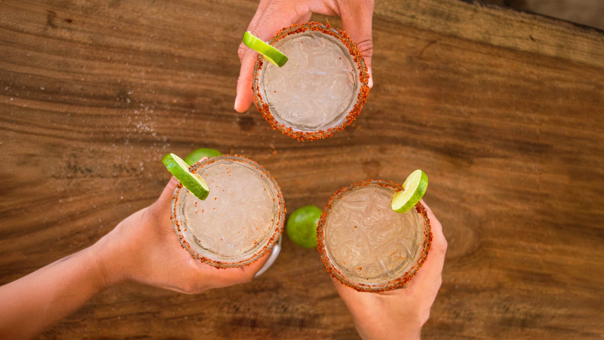 Three hands toasting with cocktail glasses garnished with lime and chili powder rim.