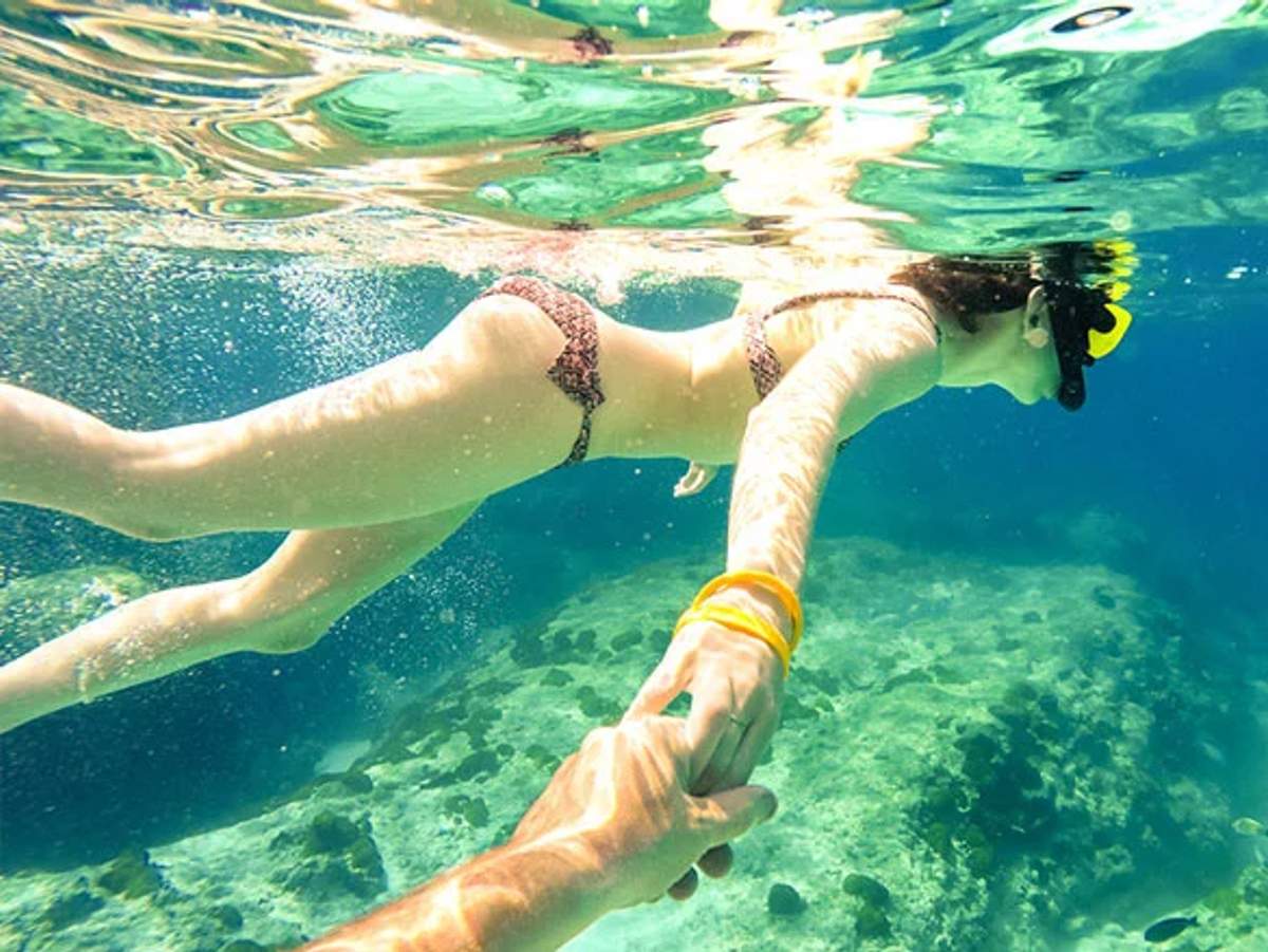 Vista submarina de una persona haciendo snorkel en agua azul clara, sosteniendo la mano de otra persona. La nadadora lleva un bikini y una máscara de snorkel, explorando el arrecife de coral debajo. La escena captura un sentido de aventura y compañerismo en el océano.