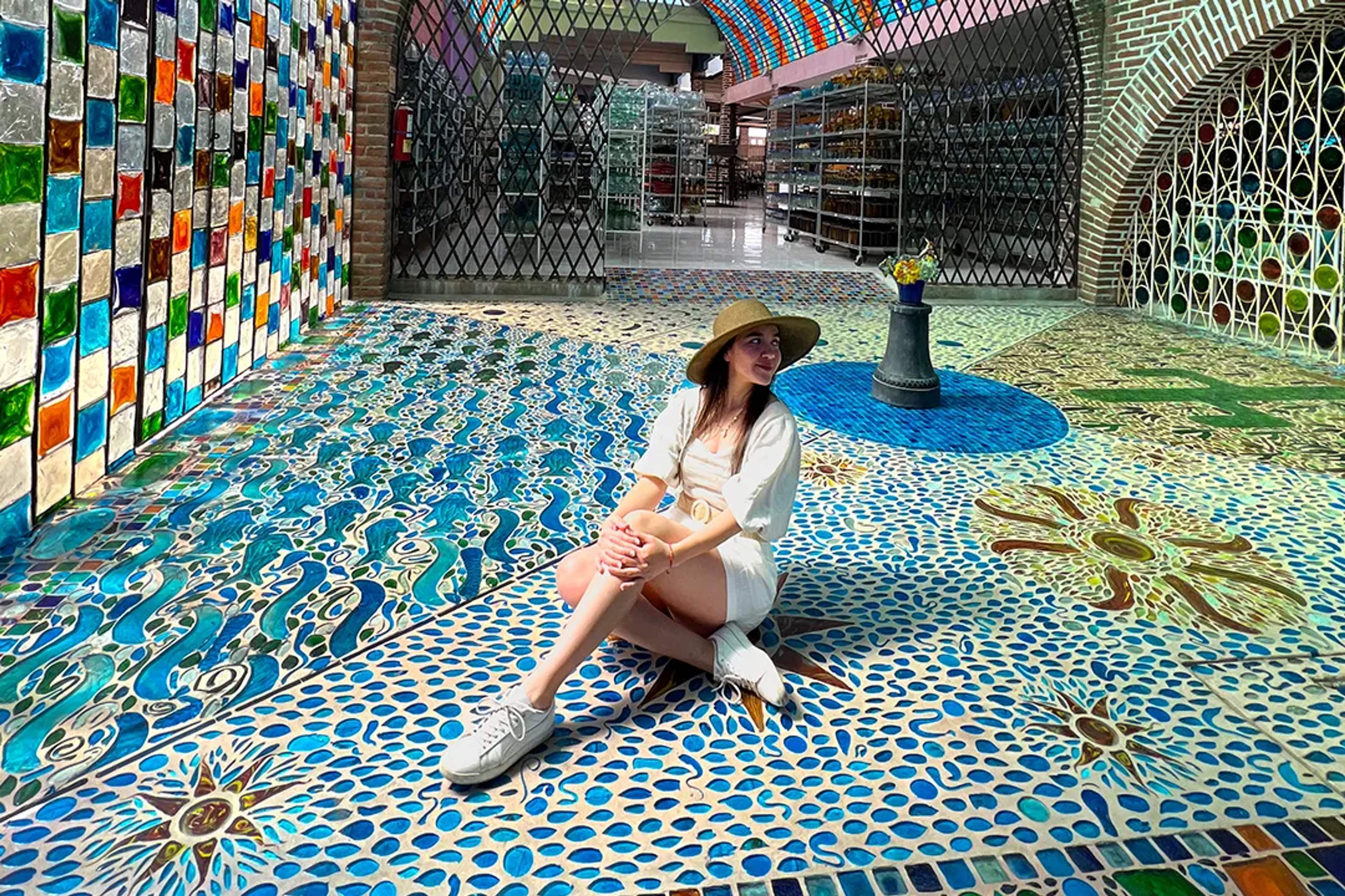 Mujer sonriente sentada sobre un piso de mosaico vibrante, rodeada de vidrio y arte colorido.