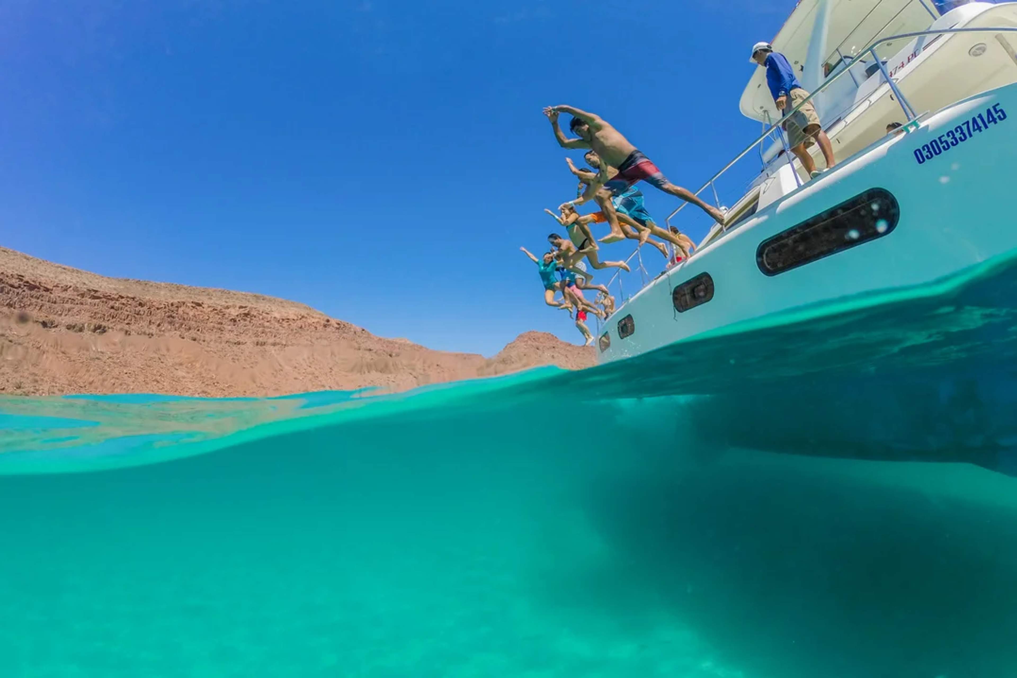 Group of friends jumps off a yacht into Balandra's clear turquoise water on a sunny day.