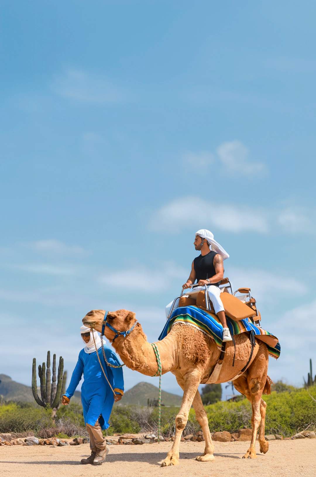 Man riding a camel led by a local guide in a desert landscape with cacti