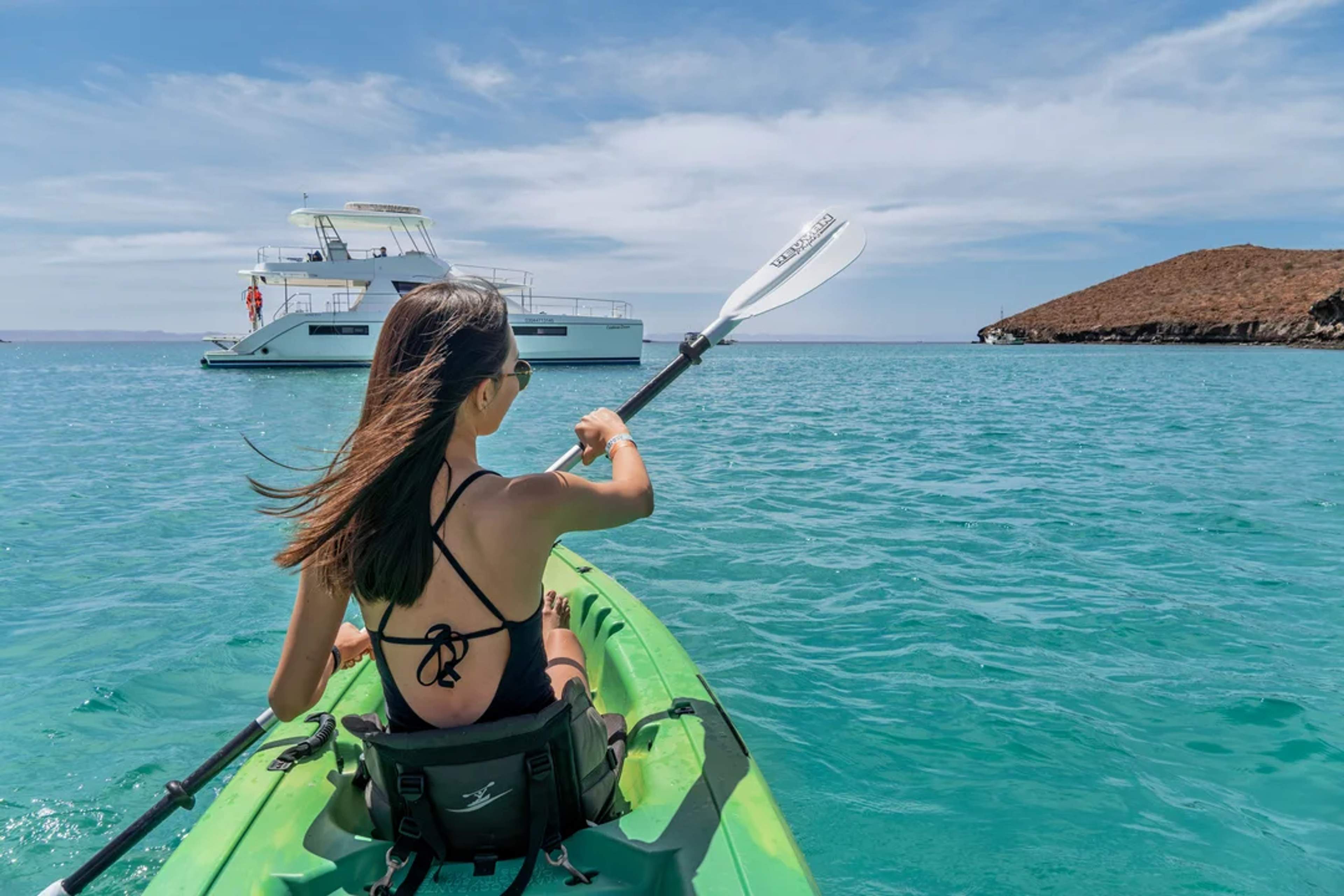 Una mujer rema en kayak en aguas cristalinas, con un yate de lujo y una isla escénica en el fondo.