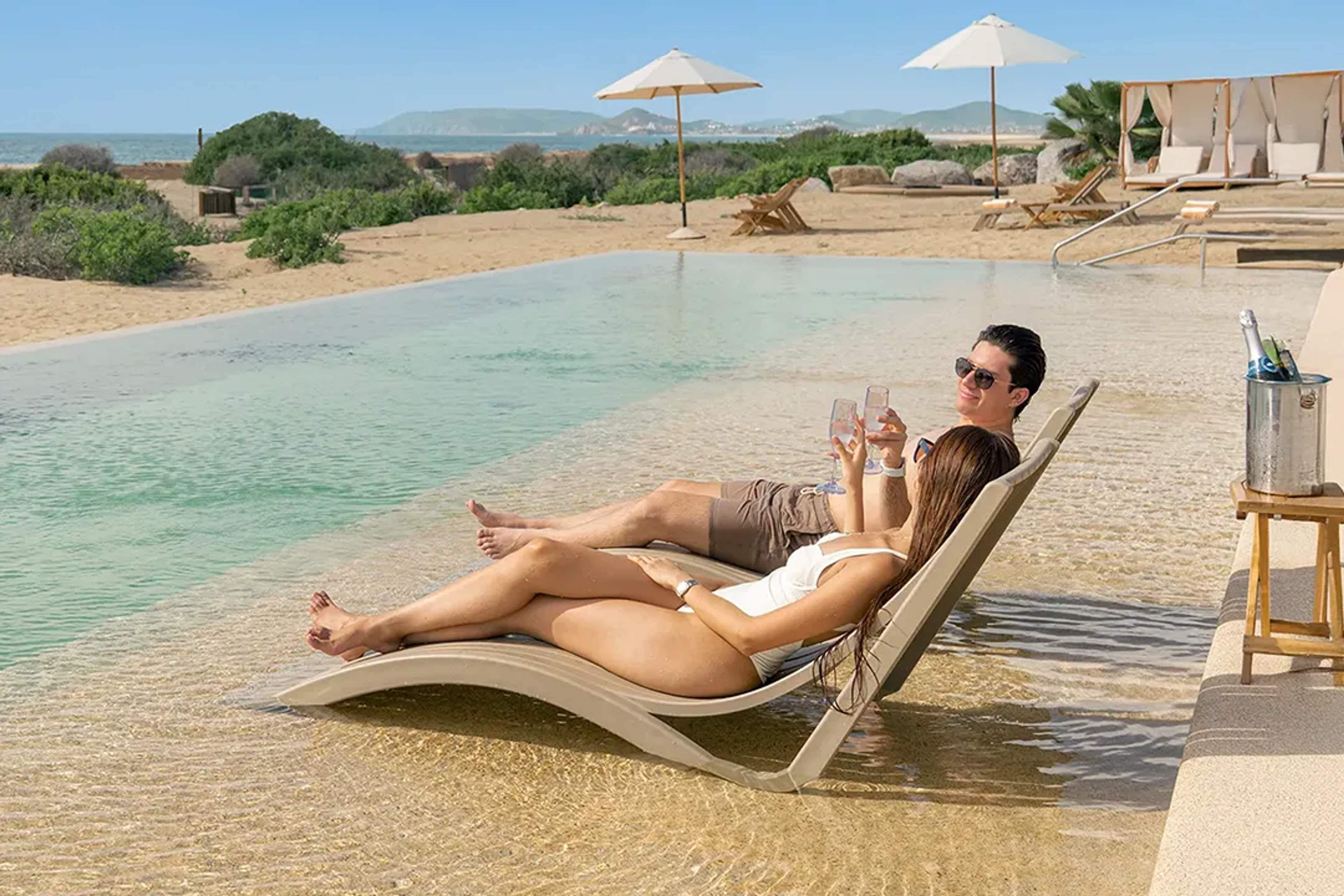 Pareja relajándose con champaña en camastros dentro de alberca infinita en Tierra Sagrada Cabo