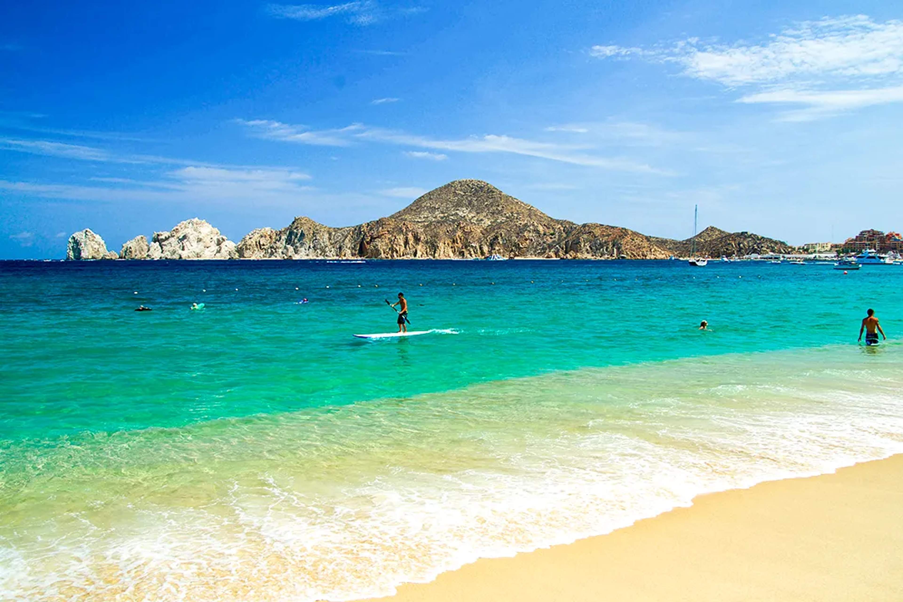 Paddleboarder on Medano Beach in Cabo San Lucas with turquoise waters and Land's End in the background.