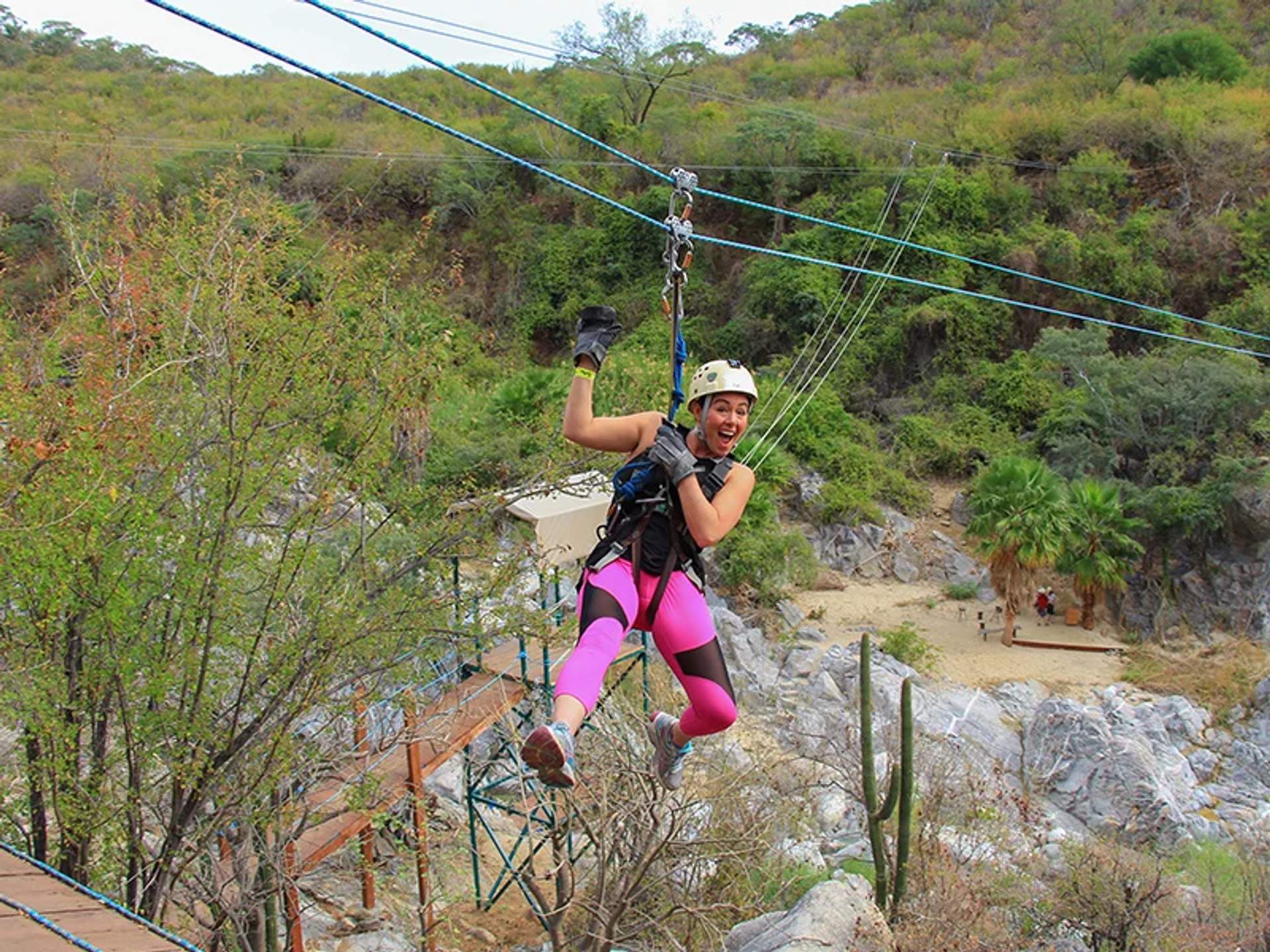 Una mujer con casco y arnés disfruta de una tirolesa sobre un paisaje rocoso y pintoresco con vegetación.
