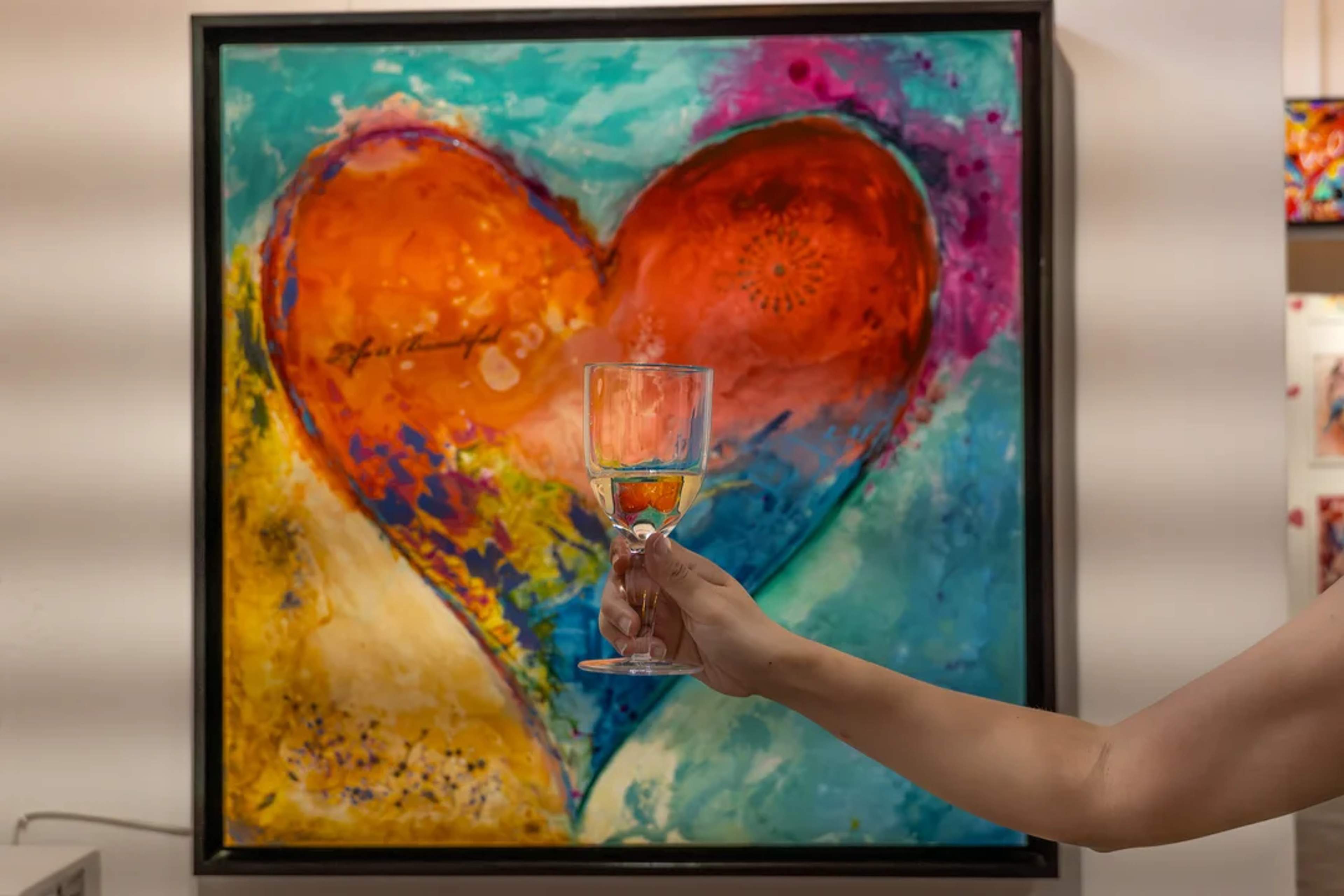 Una mano sostiene una copa de vino frente a una pintura vibrante de corazón, fusionando arte y vino en una galería colorida.