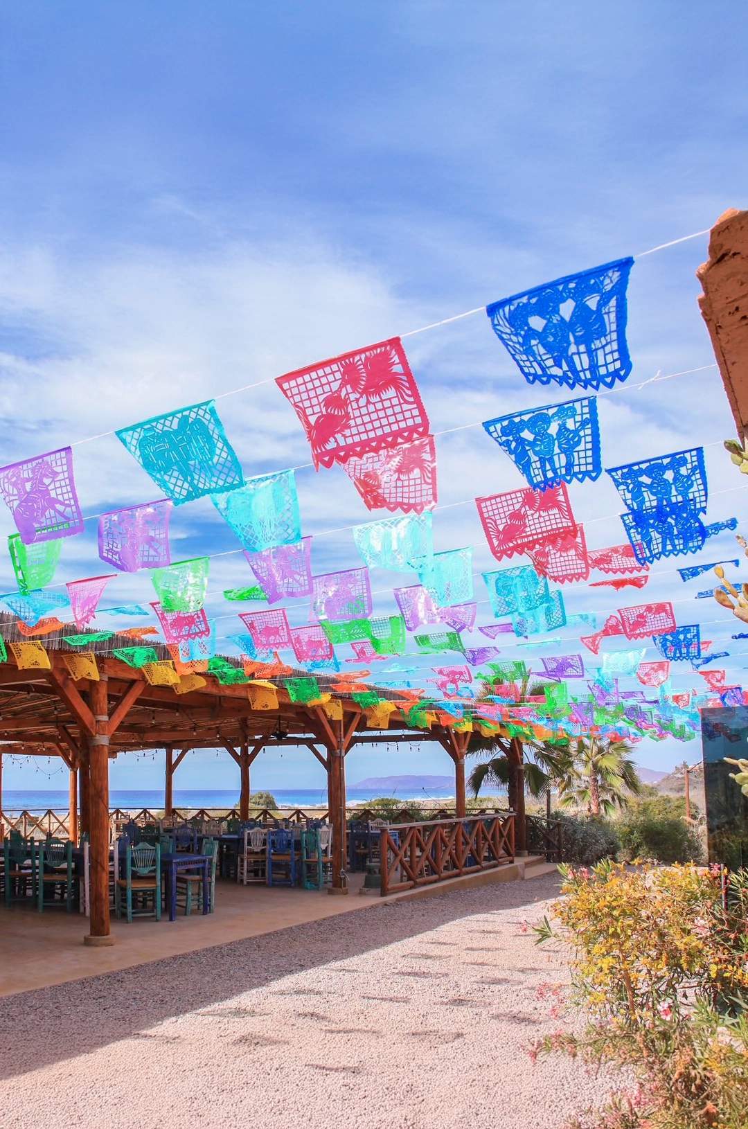 Área de comedor al aire libre decorada con coloridos banderines de papel picado, con vista al océano bajo un cielo azul.