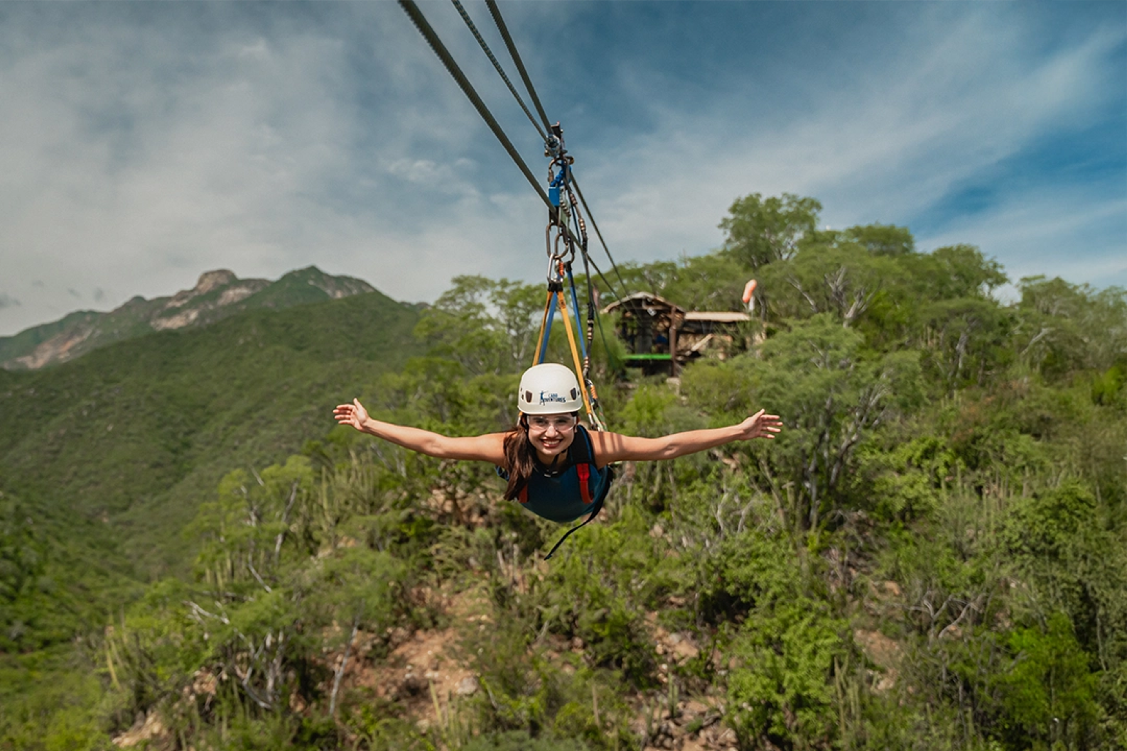 Woman smiling as she glides on a zipline over a mountainous landscape filled with greenery.