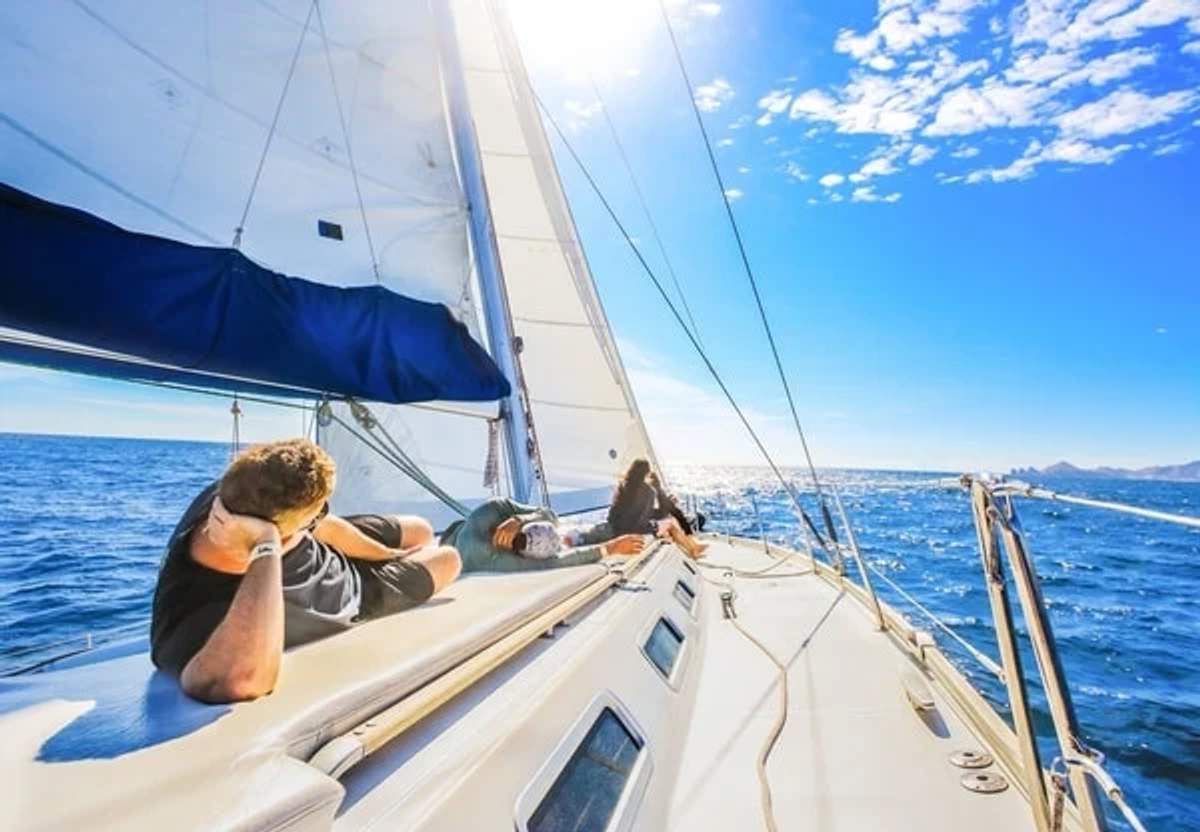 Two people relax on the deck of a sailboat under a bright, sunny sky, with the boat cutting through the calm blue ocean. The sails are fully extended, and the horizon stretches out in the distance, creating a serene and picturesque sailing scene.