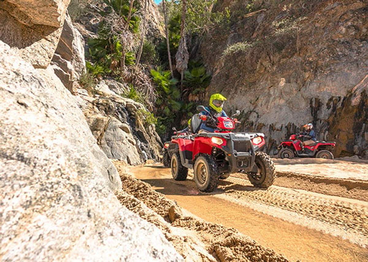 Two riders on red ATVs navigating a sandy, rocky trail surrounded by cliffs and lush vegetation in Cabo.