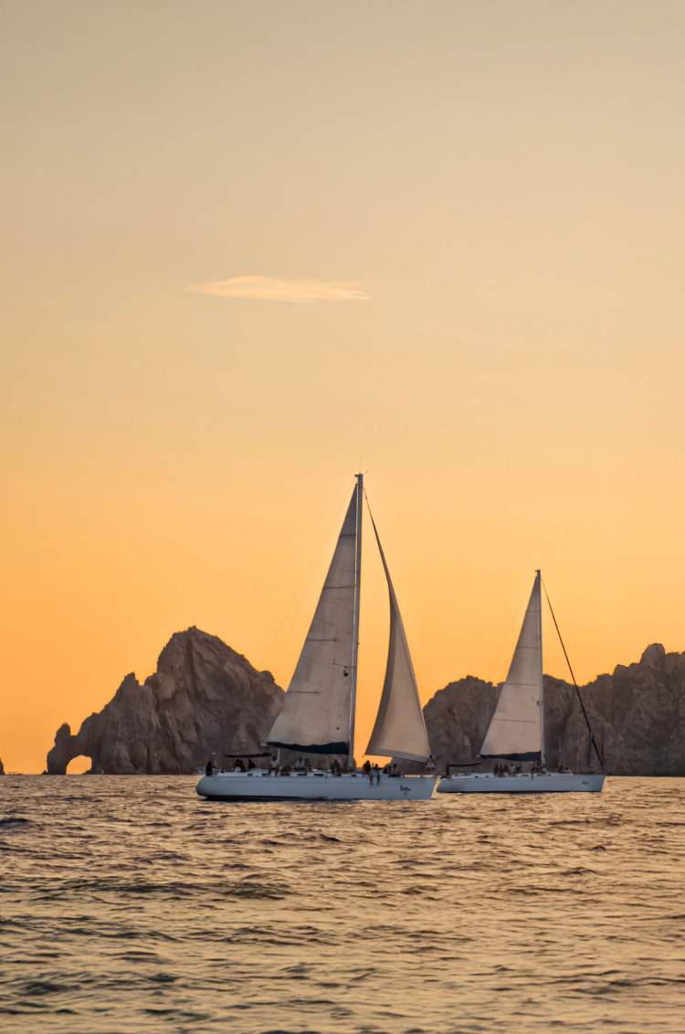Two sailboats cruise near Cabo’s Arch at sunset, with a warm orange sky and iconic rock formations in the background.