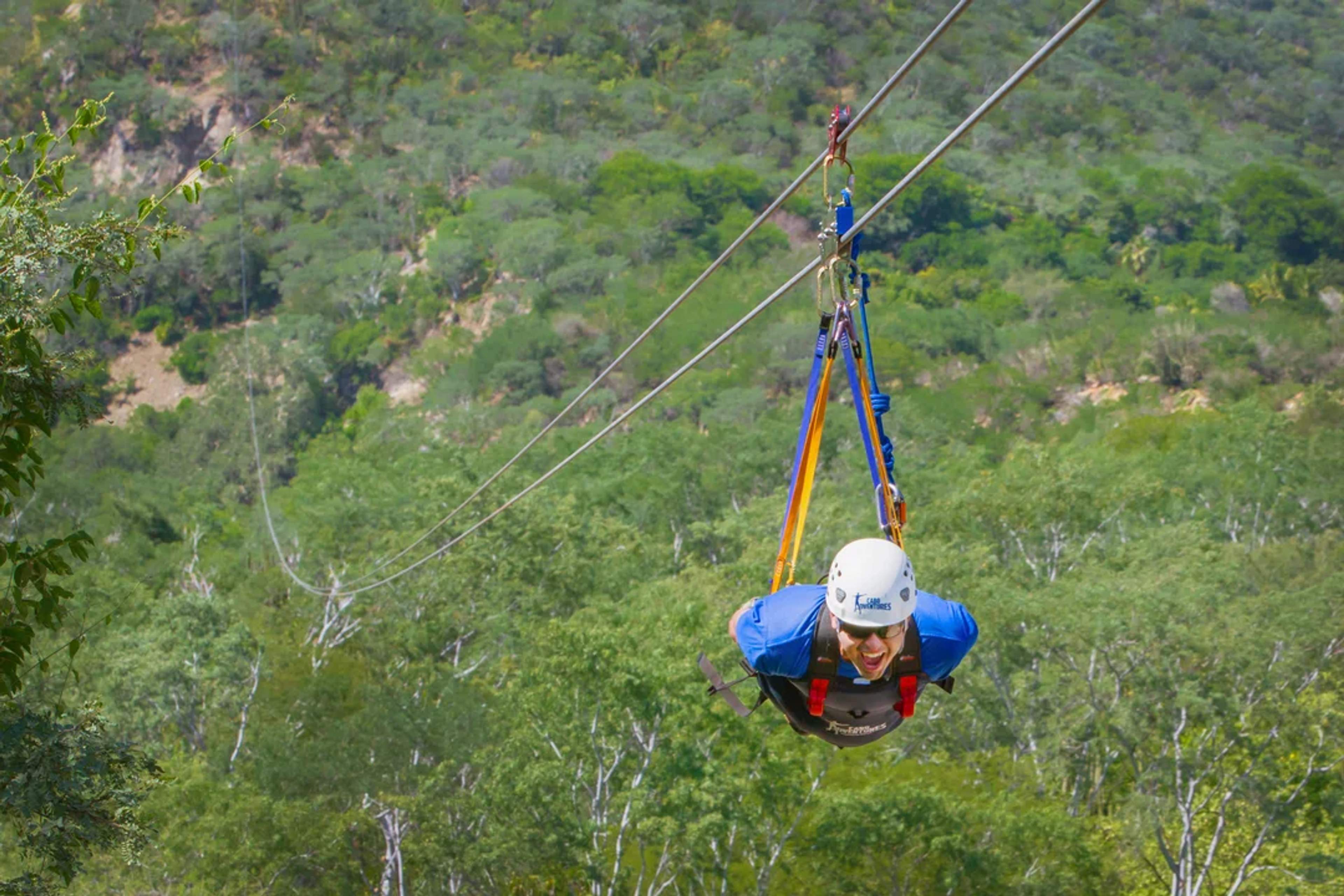 Hombre vuela estilo Superman en tirolesa sobre un cañón verde de Cabo, sonriendo con emoción.