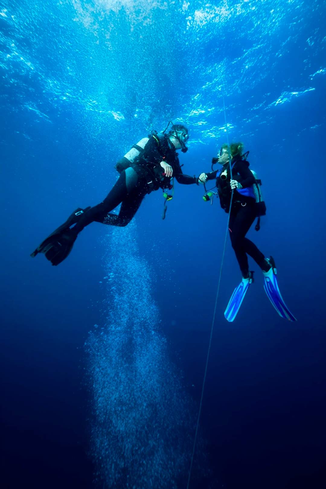 Two scuba divers hold onto a guide rope underwater, communicating as bubbles rise around them in the deep blue ocean.