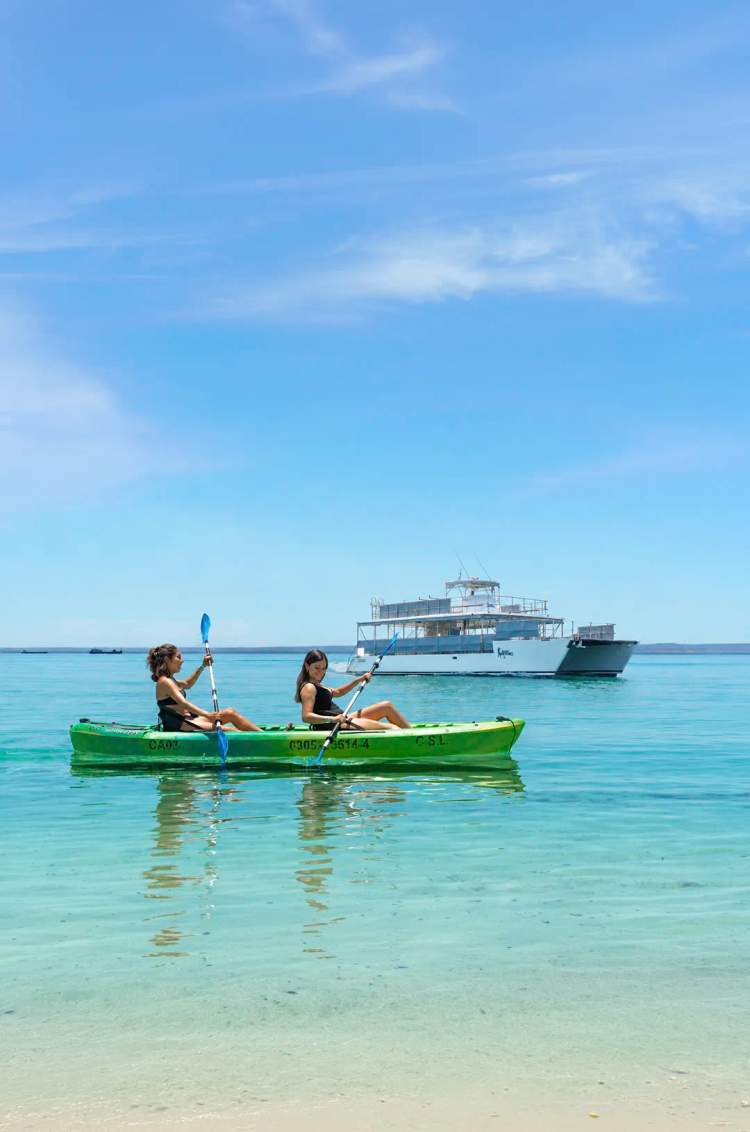 Dos personas en kayak sobre aguas turquesa de Balandra con un catamarán al fondo.