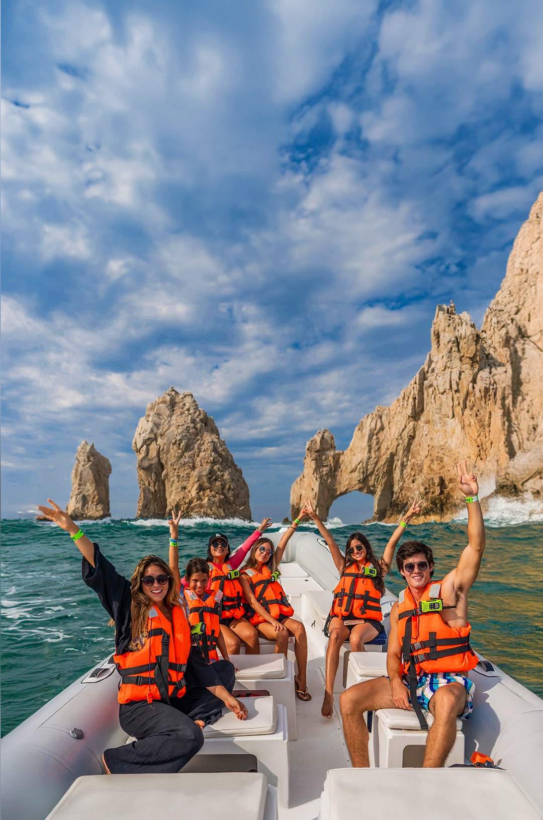 Group of people on a boat in front of the Arch of Cabo San Lucas, waving and having fun