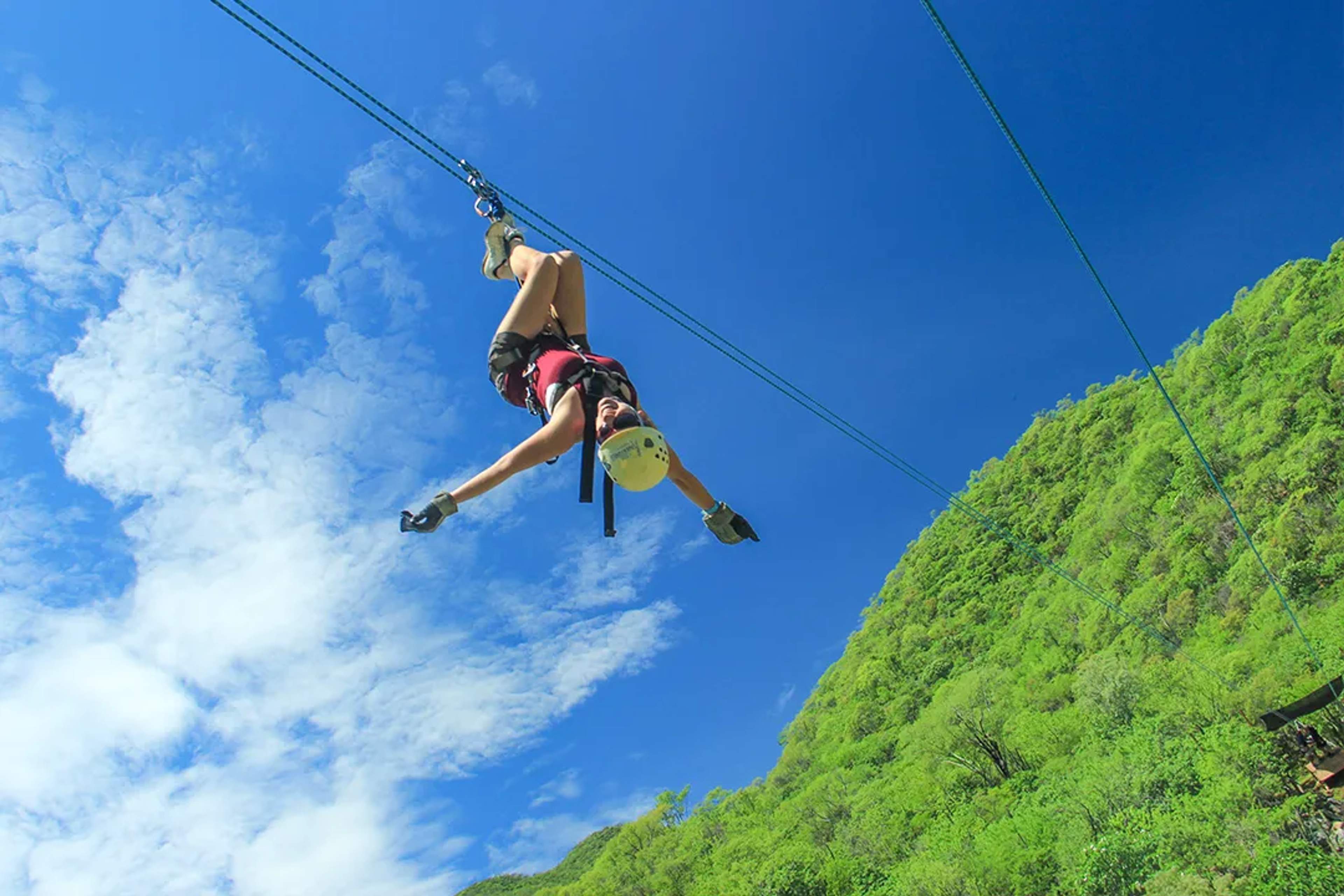 Woman ziplining upside down over lush green mountains under a bright blue sky.