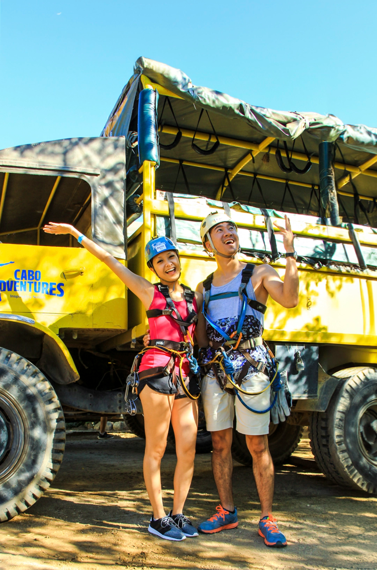 Una pareja sonriente con equipo de aventura posando frente a un camión amarillo de Cabo Adventures, con cascos y arneses.