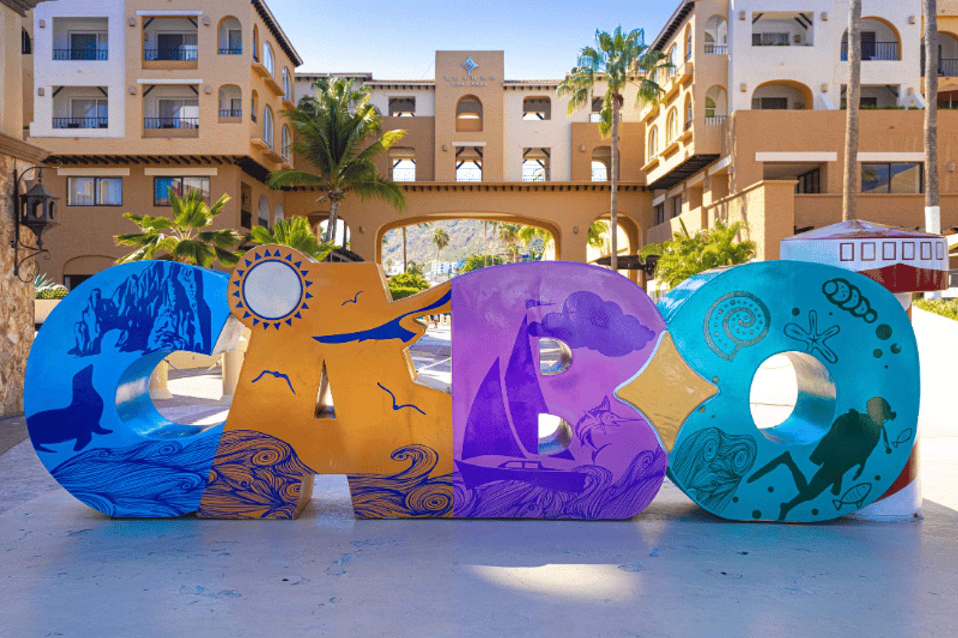 Letrero colorido de "CABO" con ilustraciones vibrantes, situado frente a un complejo turístico con palmeras y un arco de fondo.