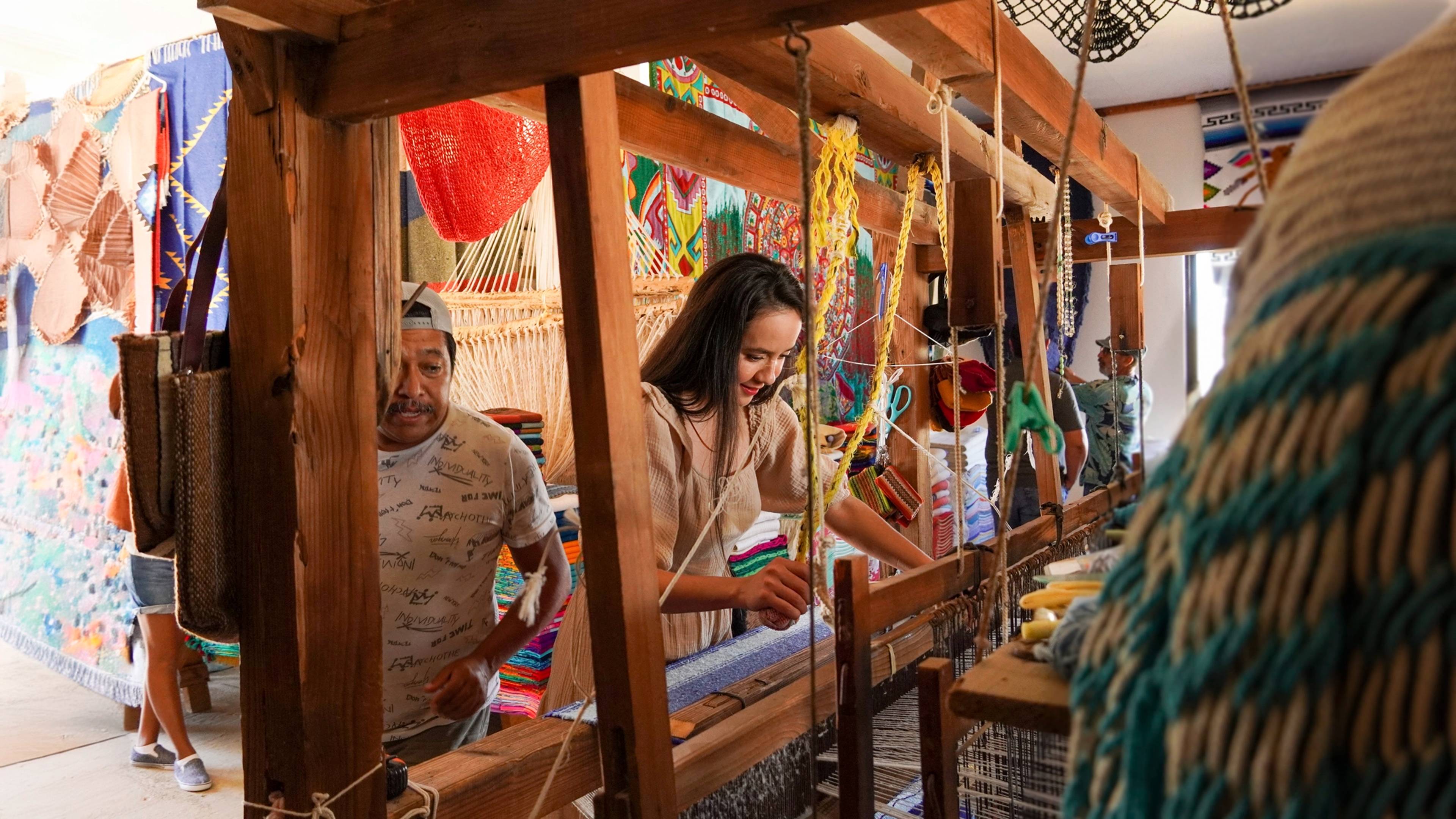 Mujer tejiendo en un telar en un mercado colorido, con un hombre cerca, rodeados de artesanías y textiles hechos a mano.