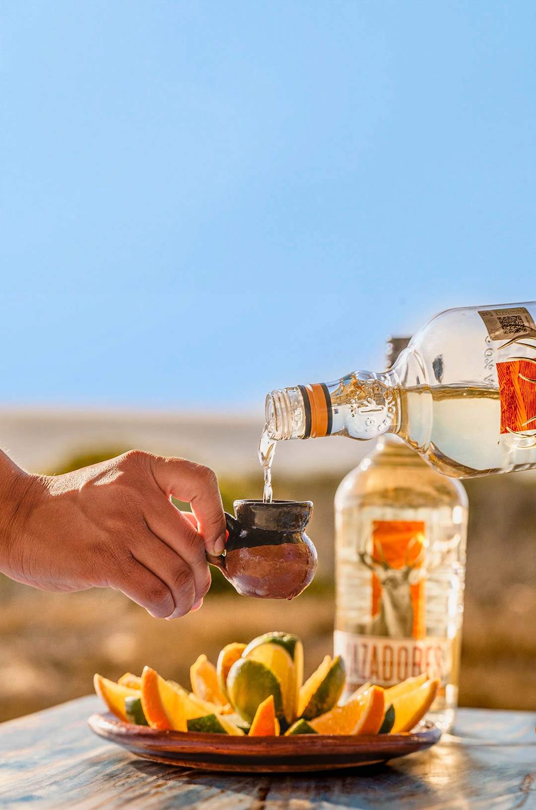 Hand pouring tequila into a small clay cup, with lemon and orange slices in the background.