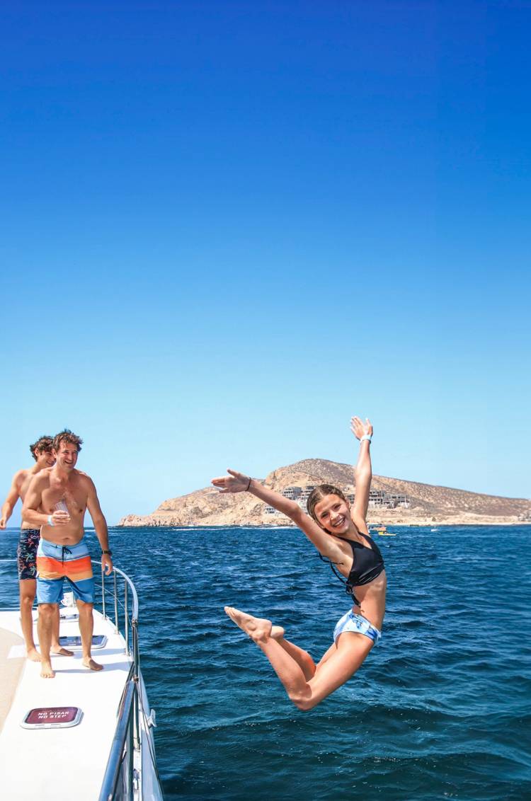 Una niña alegre salta de un barco al océano, con amigos animándola detrás, bajo un cielo azul claro.