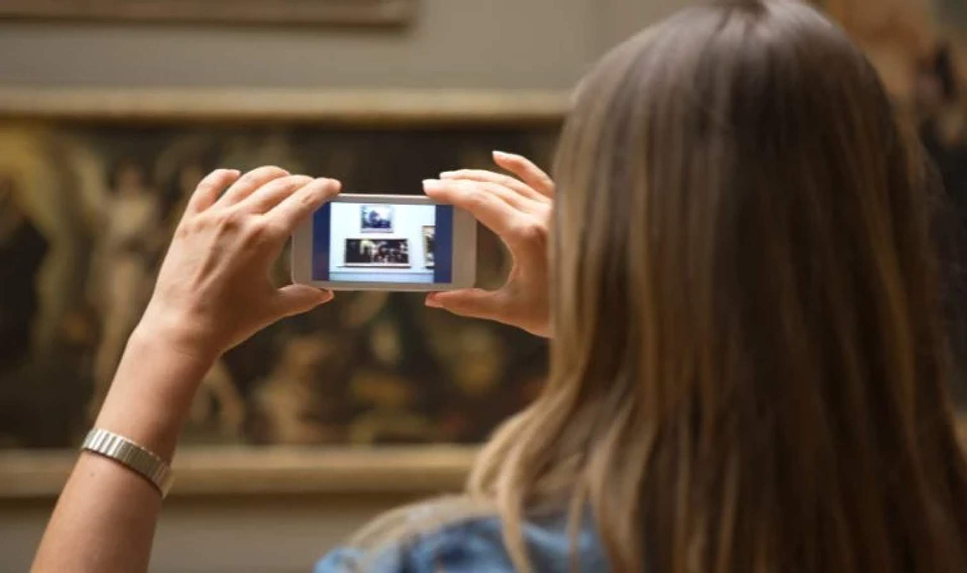 A woman taking photos of a museum display with her smartphone, capturing the artwork and details for later viewing.